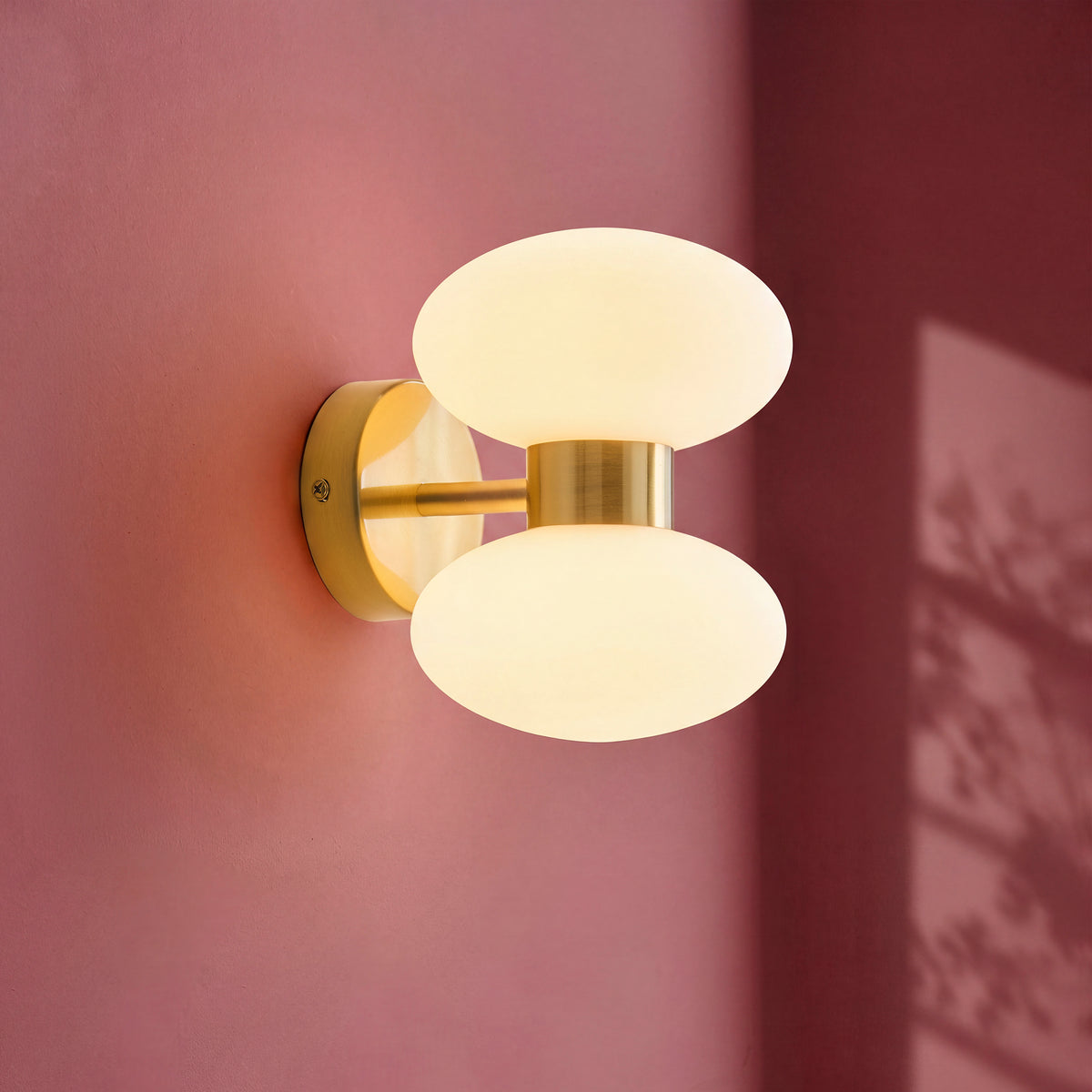 Lit lifestyle shot of the Endon Rossano 2 light wall light in warm brass with opal glass shades against a blush pink wall with dappled leaf shadow