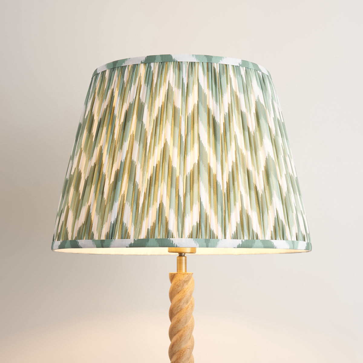 Close up of the lit Cotswold green ZigZag pleated faux silk shade and brass lampholder on the Endon Twist oak effect floor lamp