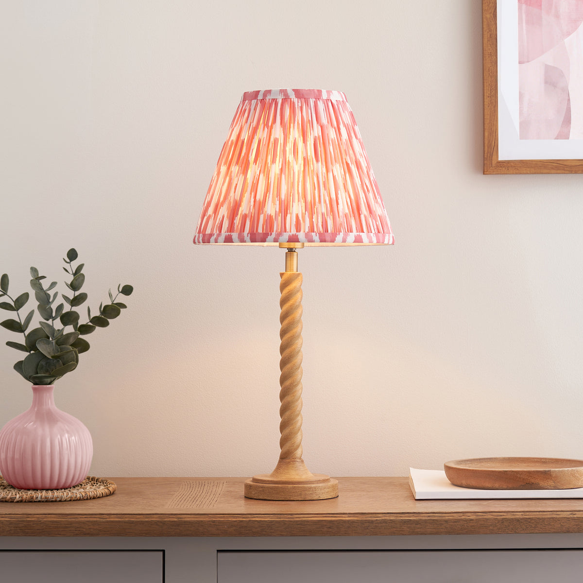 Endon Twist table lamp in oak effect resin with a coral pink ikat shade, lit, on a wooden sideboard beside a pink vase of eucalyptus and framed artwork