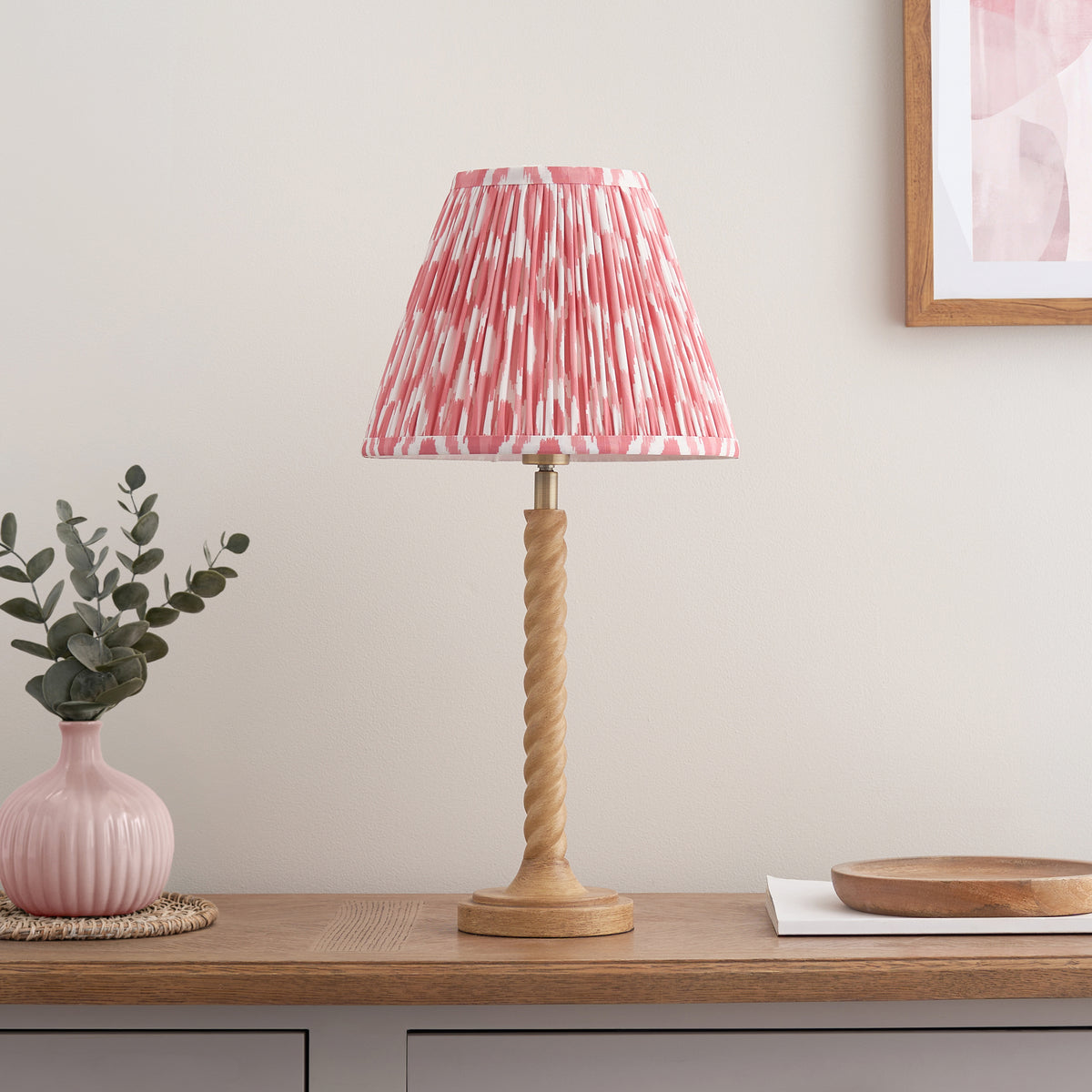 Endon Twist table lamp with oak effect twisted base and coral pink ikat shade, unlit, styled on a wooden sideboard with a pink vase and wooden bowl
