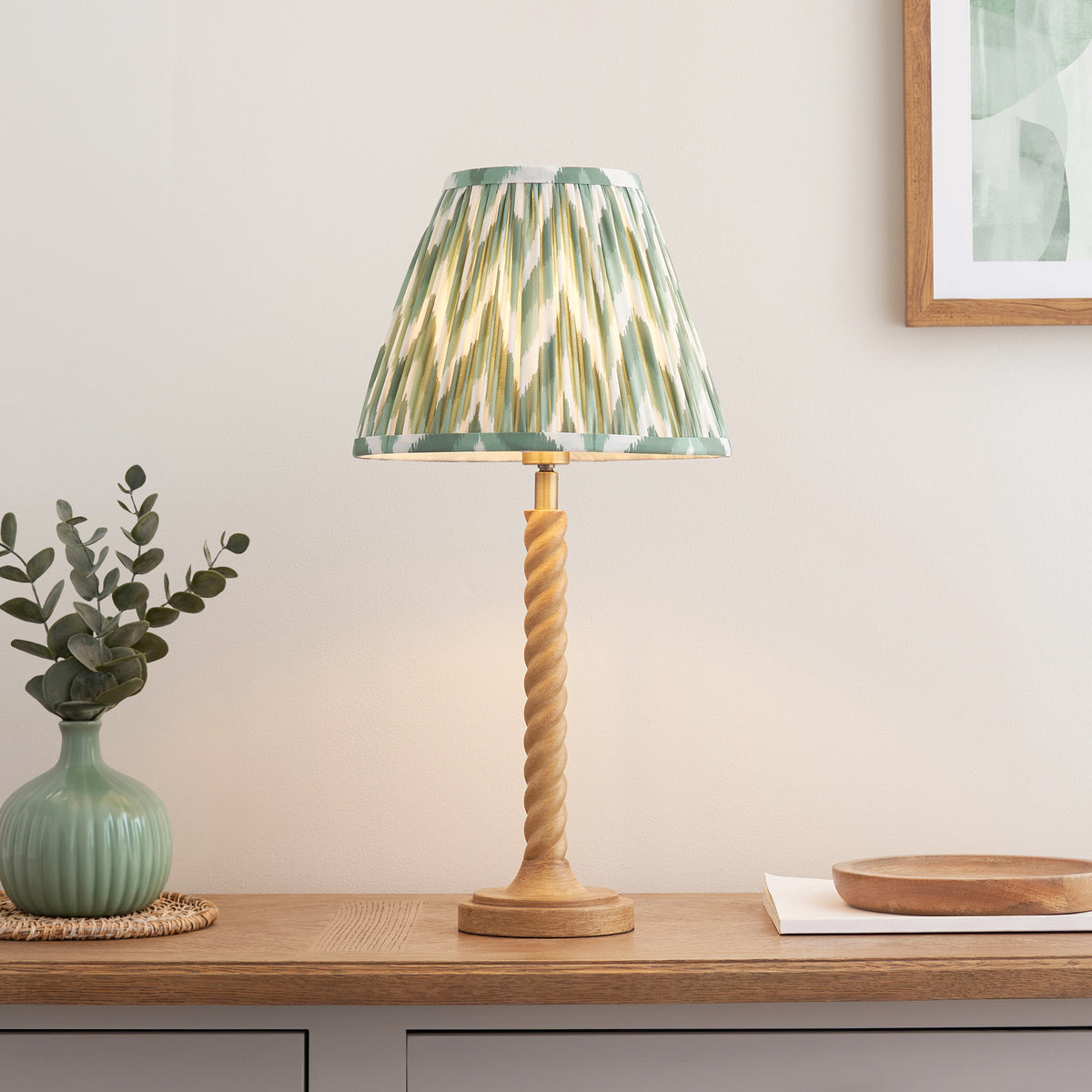 Endon Twist oak effect table lamp with green zigzag shade lit on an oak sideboard beside a sage vase, eucalyptus and a framed print