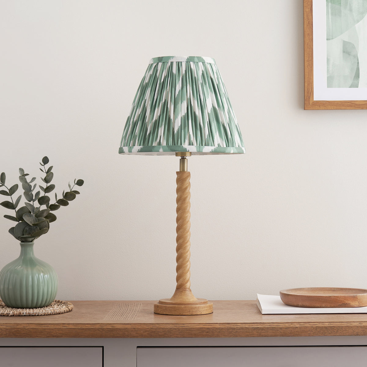 Endon Twist oak effect table lamp with Cotswold green zigzag shade switched off on an oak sideboard with a vase and framed print