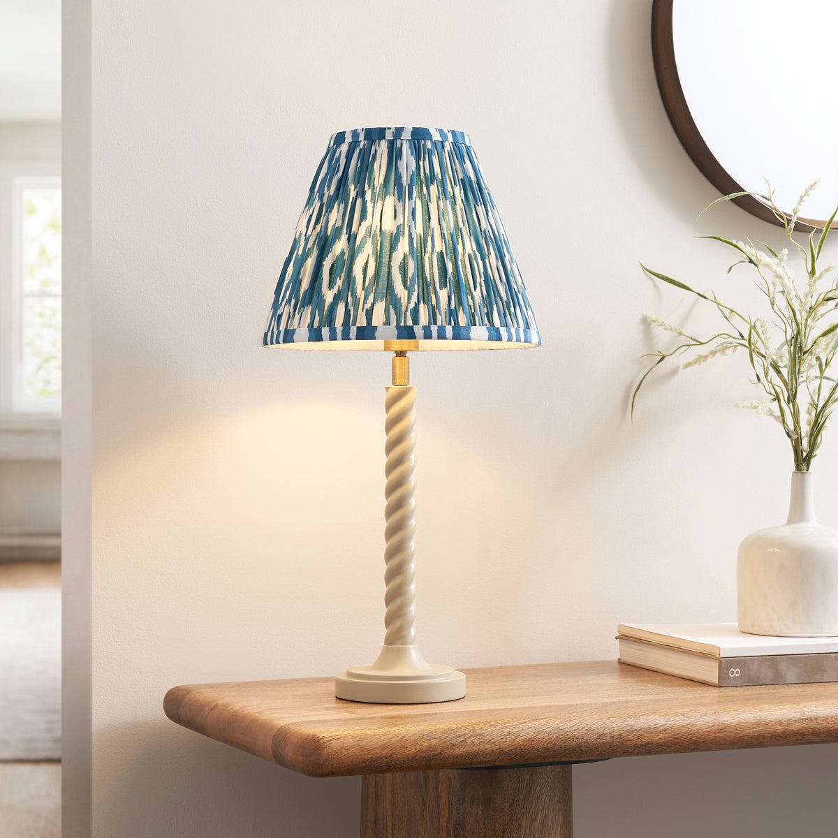 Endon Twist table lamp lit on a wooden console table beside a round mirror and a vase of dried grasses, blue ikat shade and white twisted stem