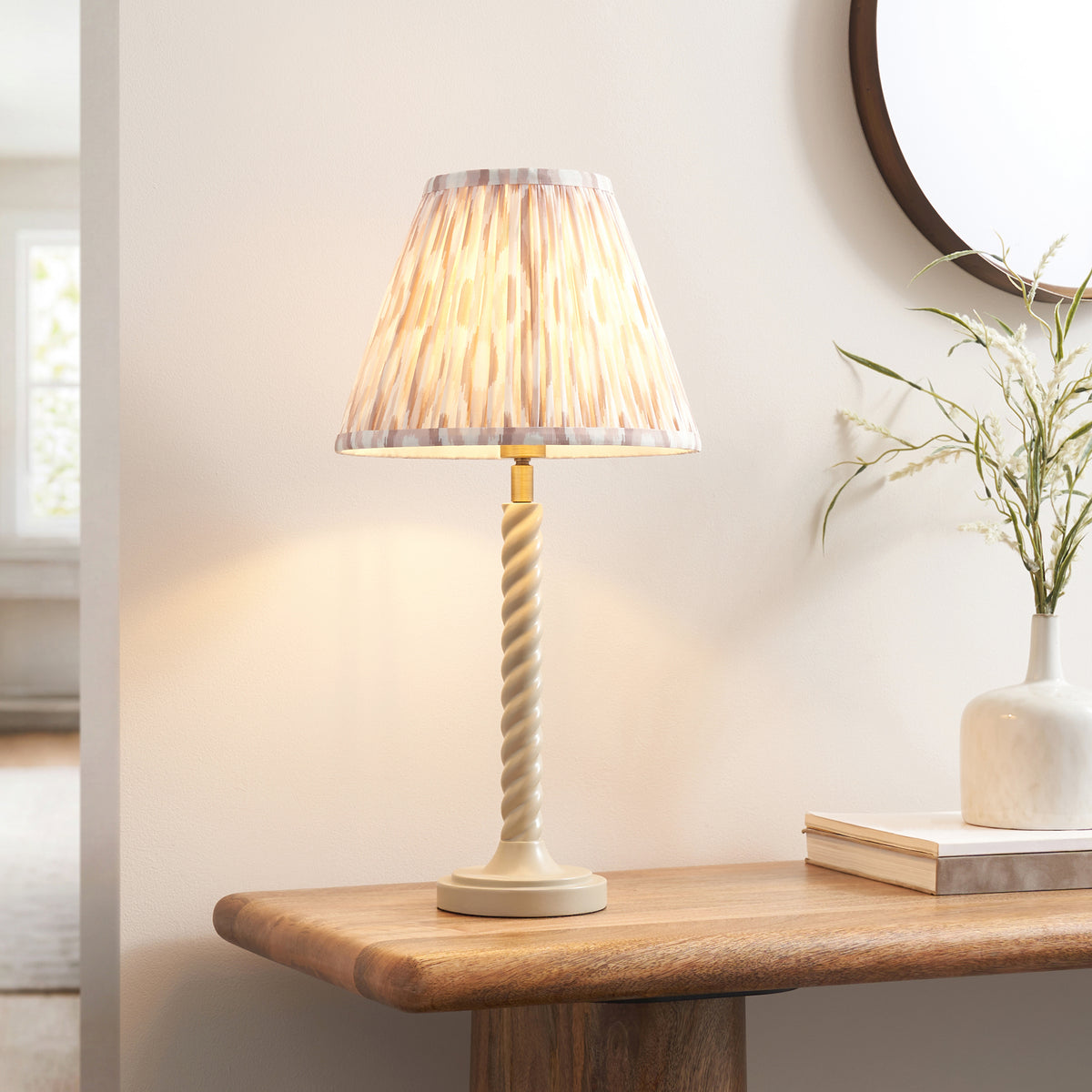 Endon Twist table lamp lit on a wooden console table beside a round mirror, white vase with dried grasses and books, almond white stem and neutral ikat shade