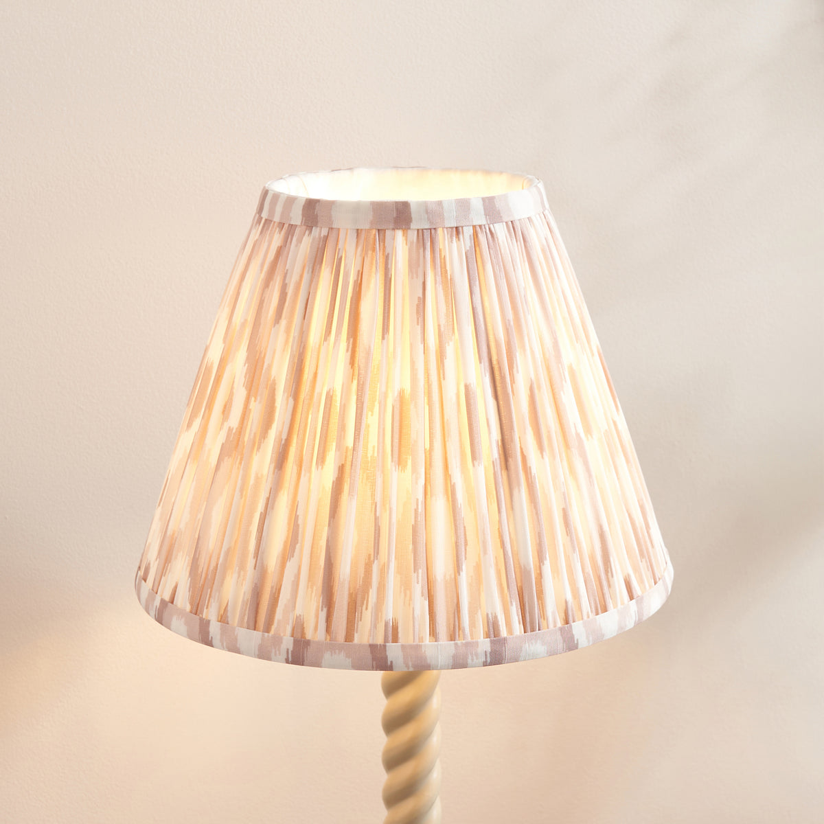 Close up of the lit neutral ikat pleated shade on the Endon Twist table lamp, blush and taupe streaks glowing against a plain wall