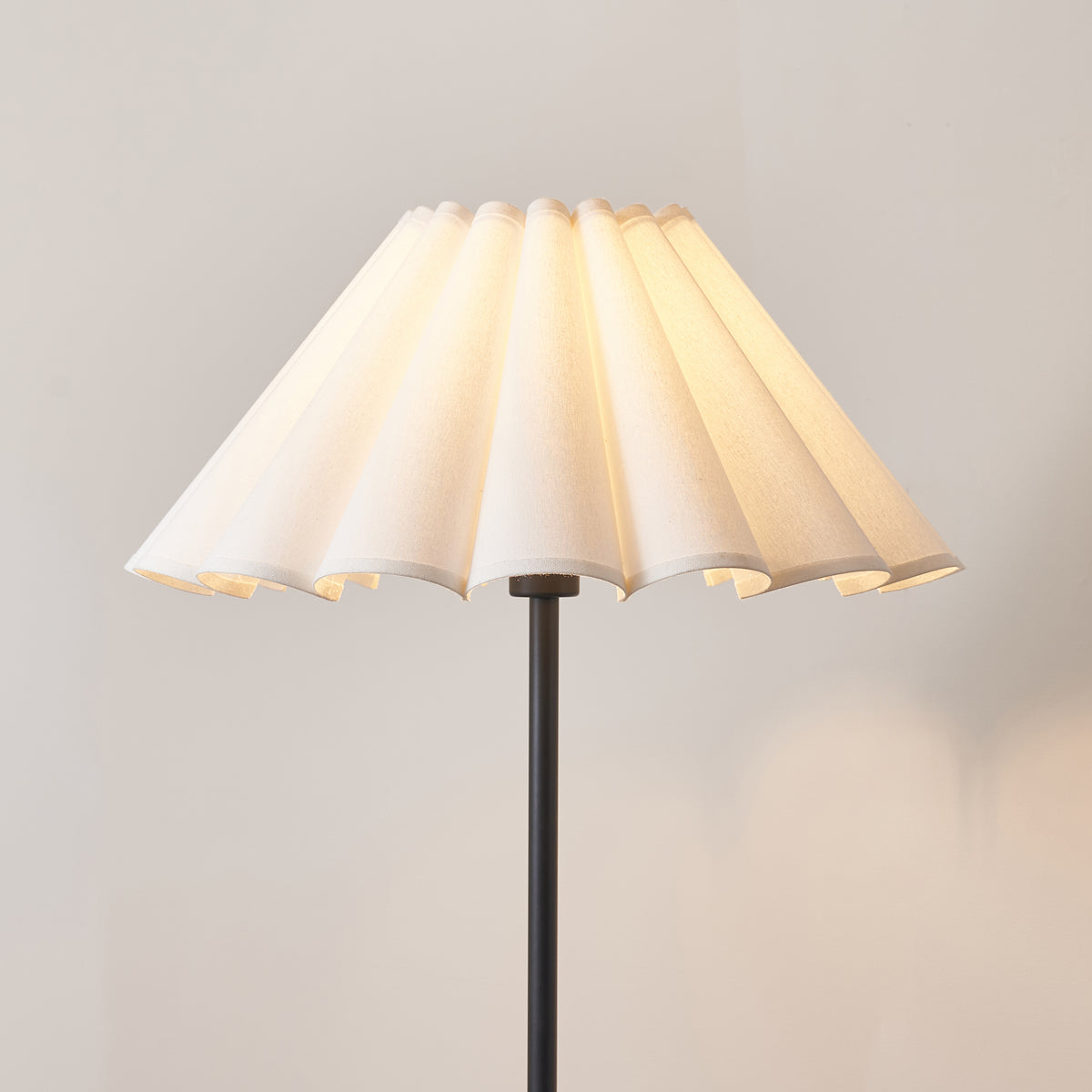 Close up of the lit Ayla 53cm scalloped cotton shade in vintage white on the Endon Arlo matt black floor lamp stem, pleats glowing cream