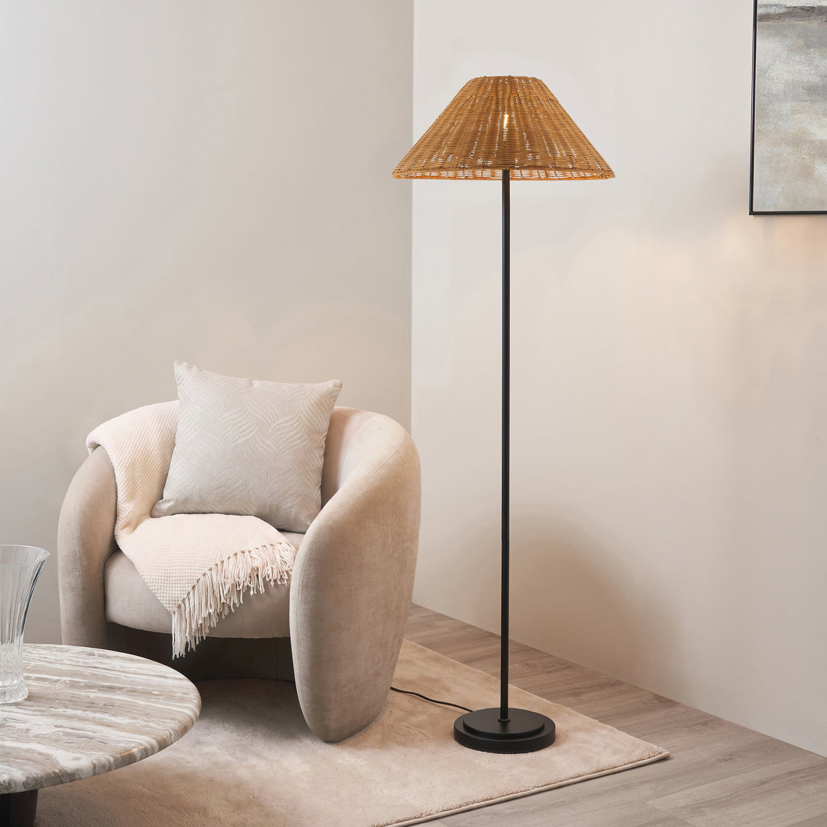 Endon Arlo floor lamp in matt black with a dark rattan shade, lit, standing beside a cream armchair in a living room