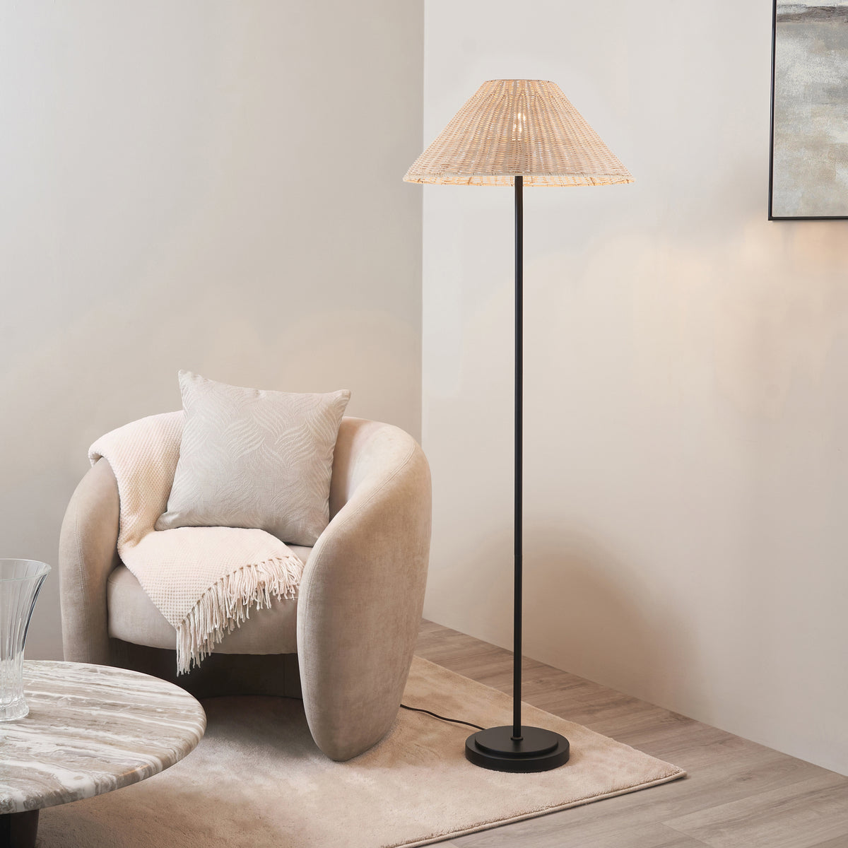Endon Arlo matt black floor lamp with natural rattan shade lit beside a beige armchair, marble table and rug in a living room