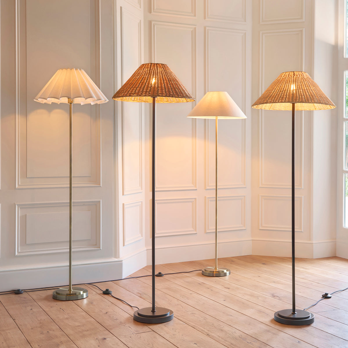 Group shot of four lit Endon Arlo floor lamps in a panelled room, including matt black stems with natural and dark rattan shades and brass stems with fabric shades