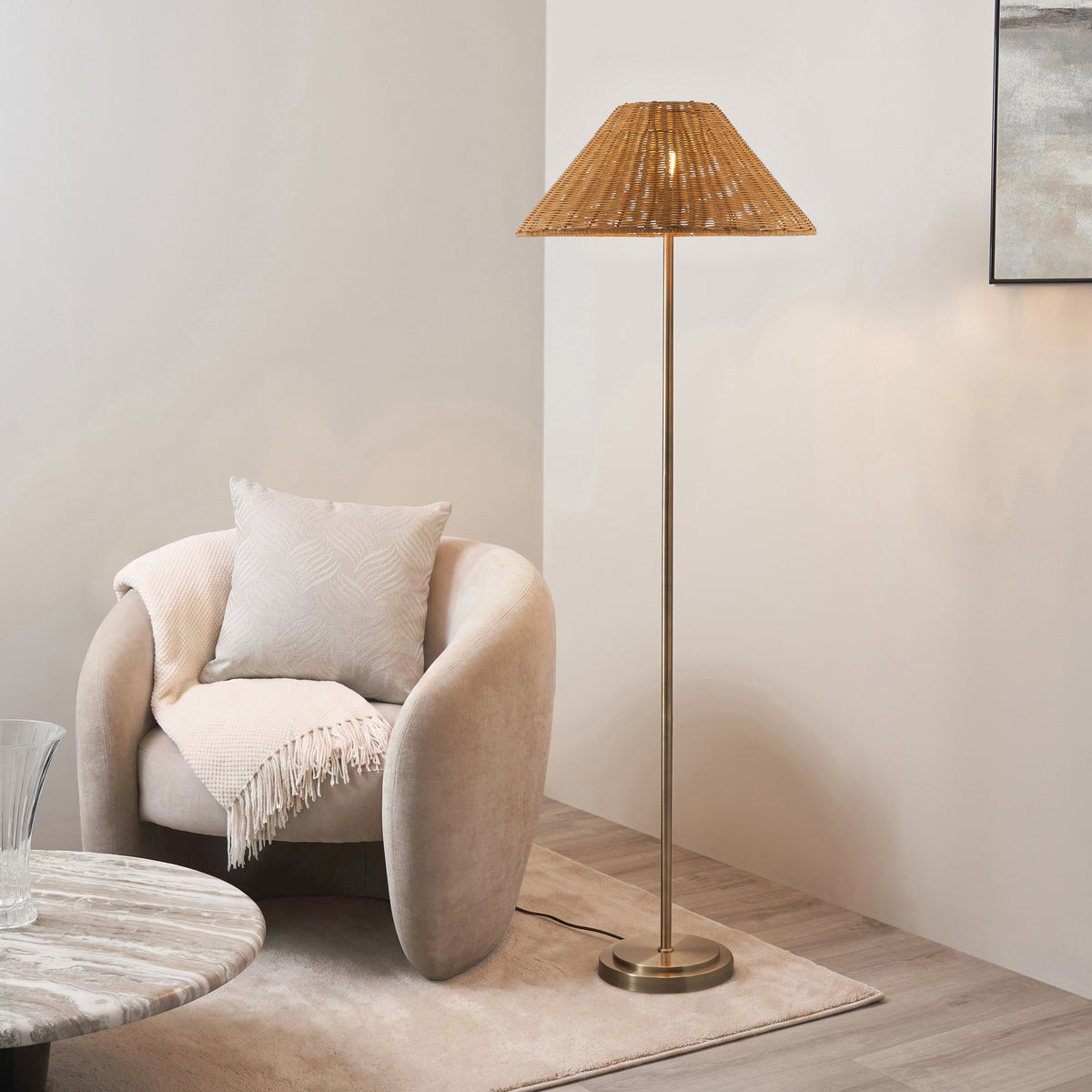 Endon Arlo antique brass floor lamp with rattan shade lit beside a cream curved armchair, marble side table and rug