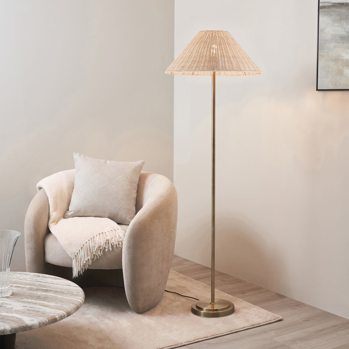Endon Arlo antique brass floor lamp with natural rattan shade lit beside a beige armchair, marble side table and rug in a living room