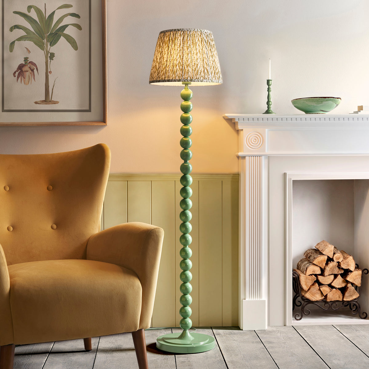 Endon Higgledy green bobbin floor lamp lit in a living room beside a mustard velvet armchair, white fireplace with logs and a botanical print