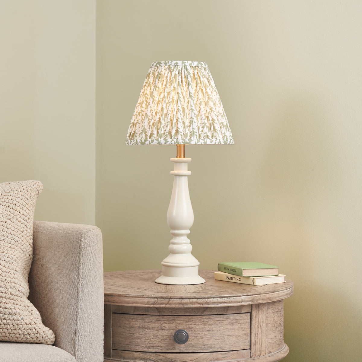Endon Myrtle table lamp lit on a round washed wood side table beside a cream sofa, green books nearby, sage green wall behind
