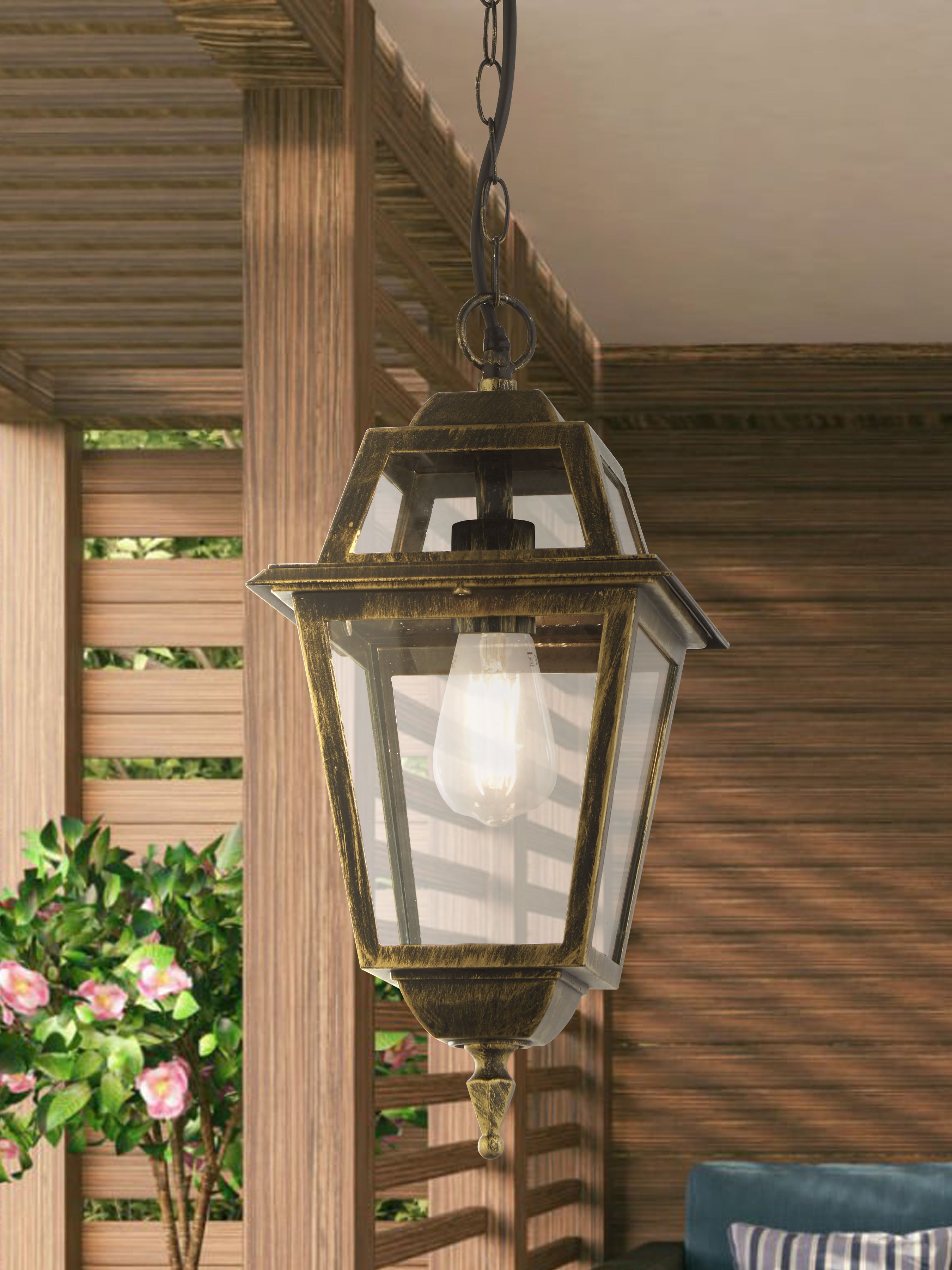 New Orleans Pendant Lantern hanging under a wooden slatted pergola on a covered patio, lit with a warm glow beside potted pink flowers and a blue armchair
