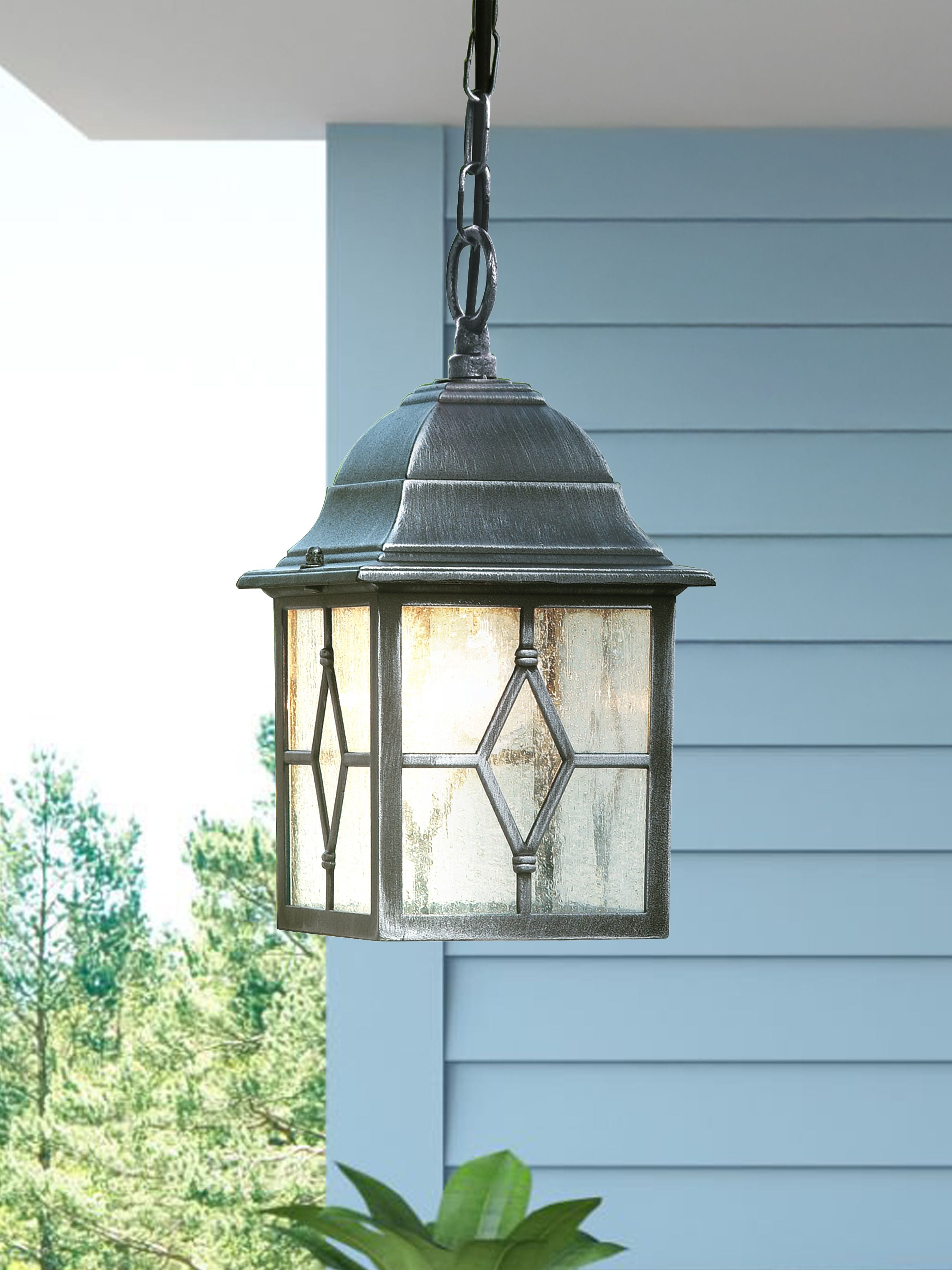 Genoa outdoor pendant hanging beneath a porch roof against pale blue clapboard siding, lit warmly at dusk with greenery in the foreground