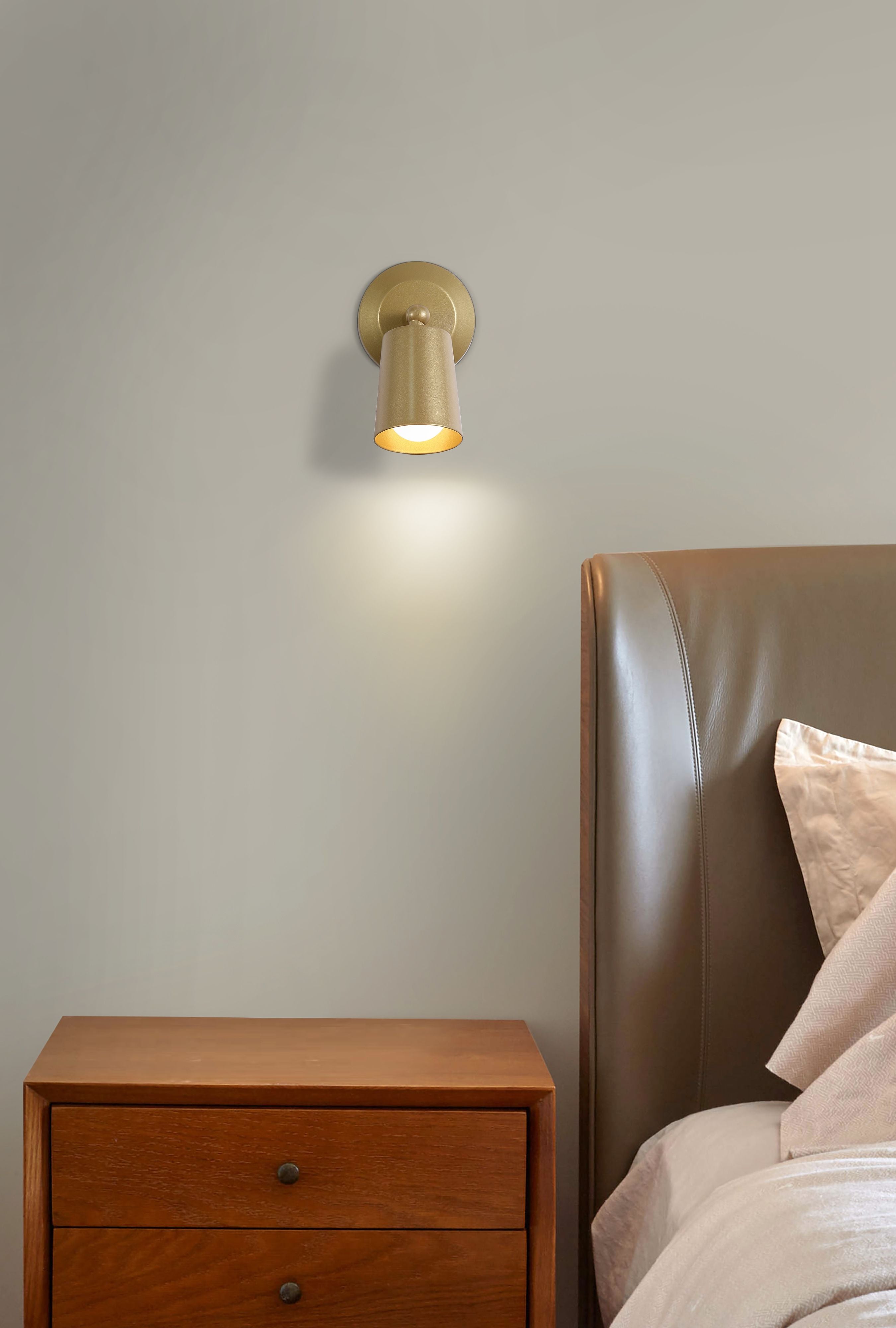 Cone gold wall light mounted above a wooden bedside table next to a brown leather headboard, casting warm light downward