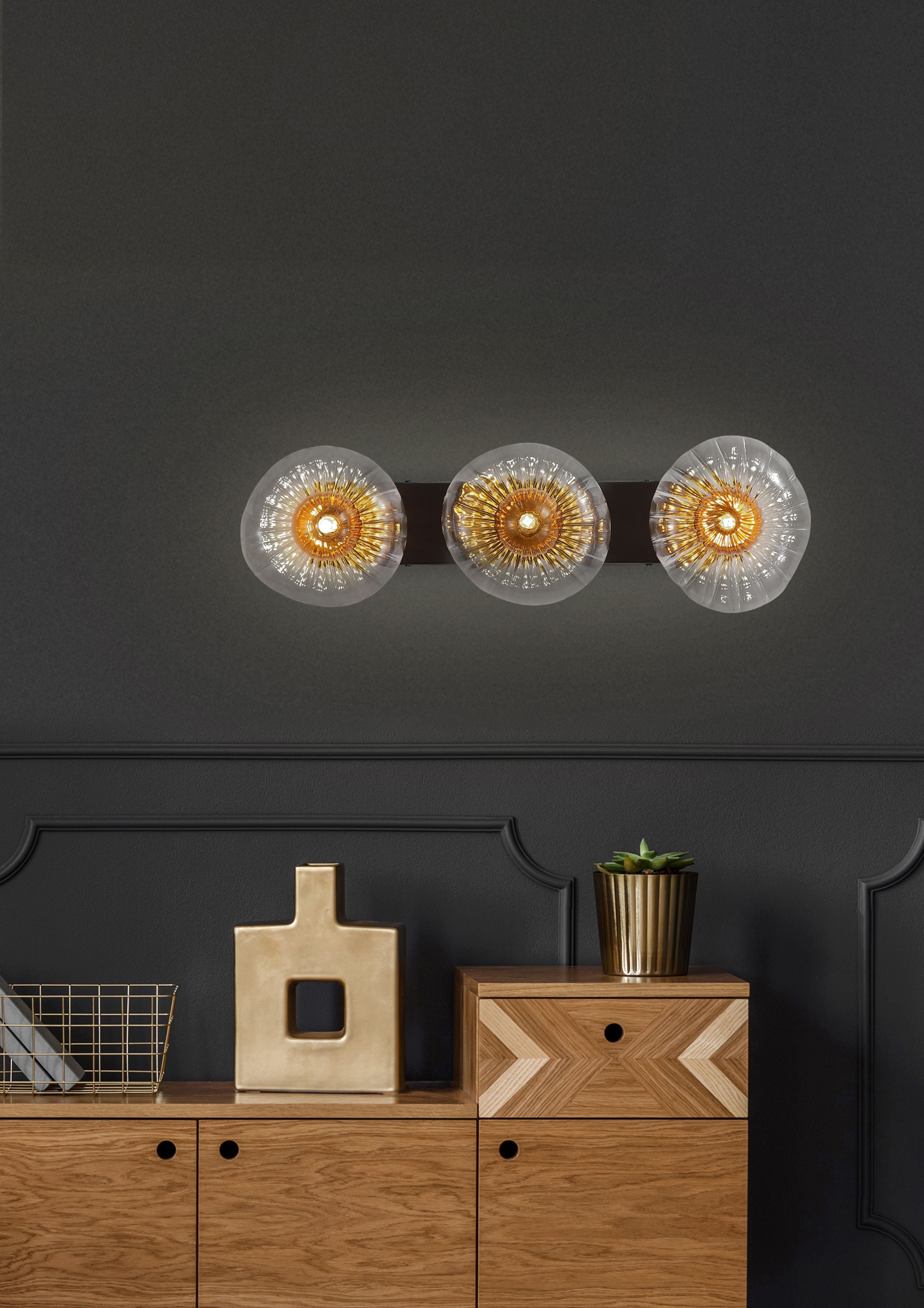 Searchlight Sunbeam 3 light wall light mounted on a dark wall above a wooden sideboard with a ceramic vase and potted plant