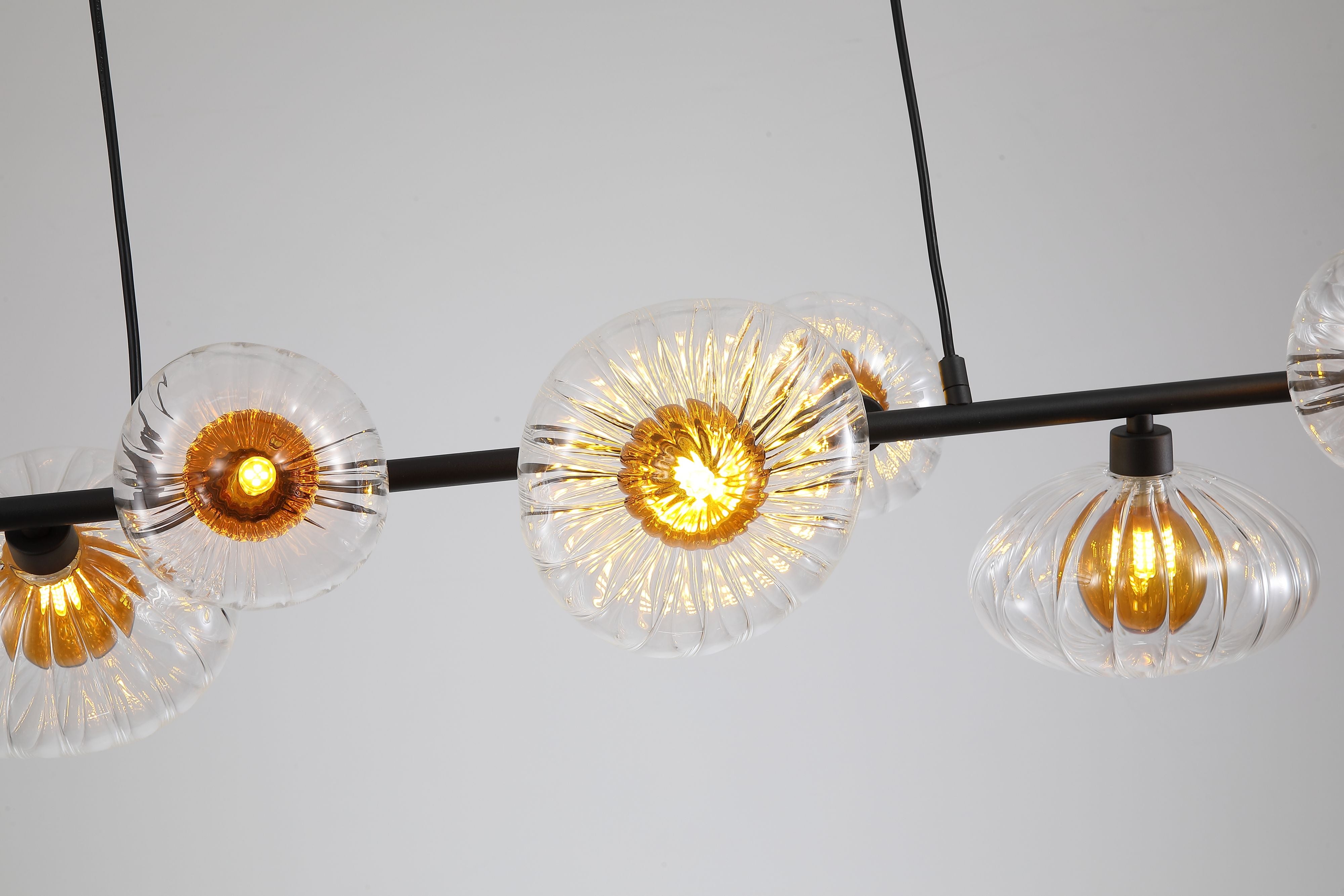 Close up of the Searchlight Sunbeam 7 light pendant showing the black bar, cable suspension and fluted glass shades glowing amber when lit