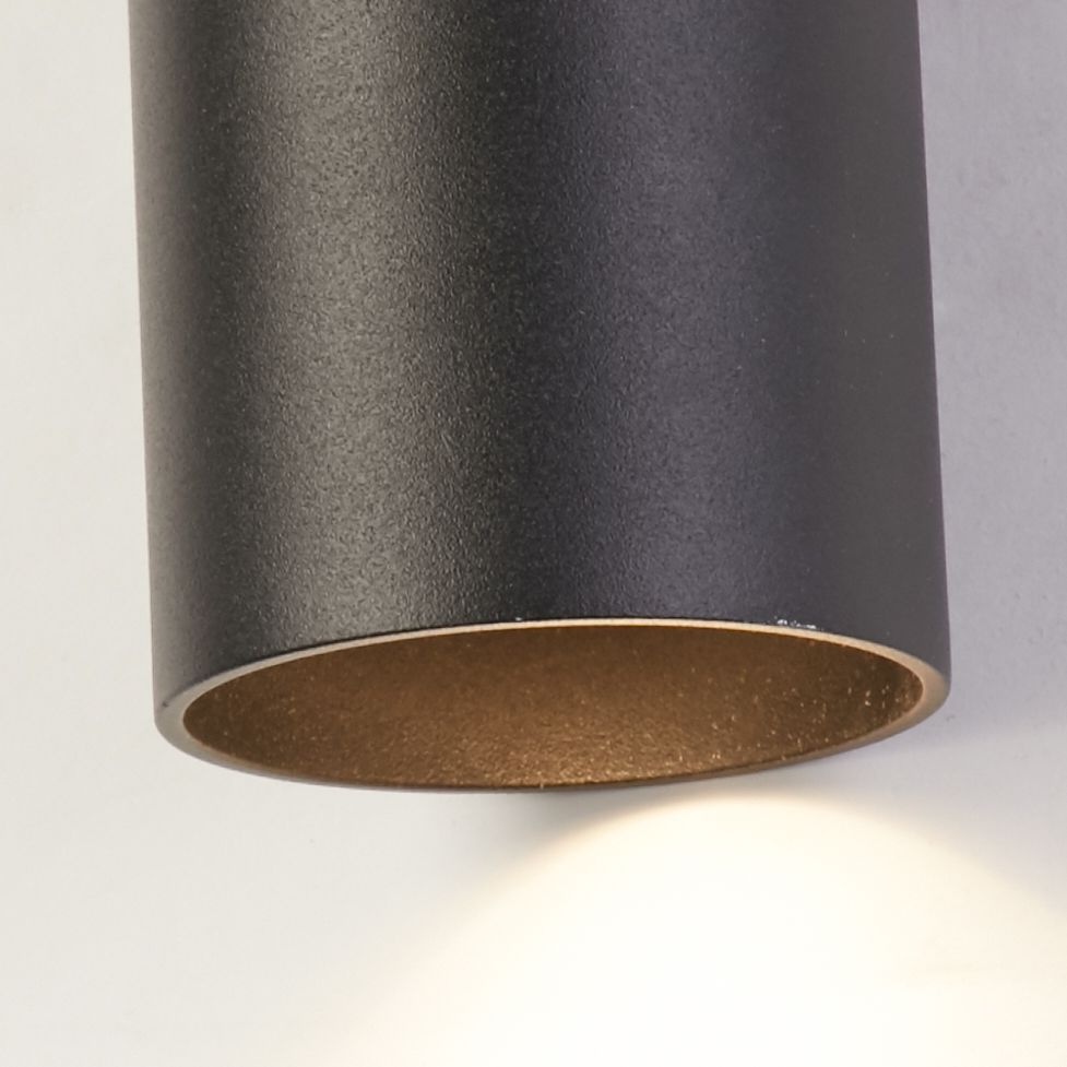 Close up of the Searchlight Cyls wall light in matte black showing the textured finish and lit lower aperture against a white wall