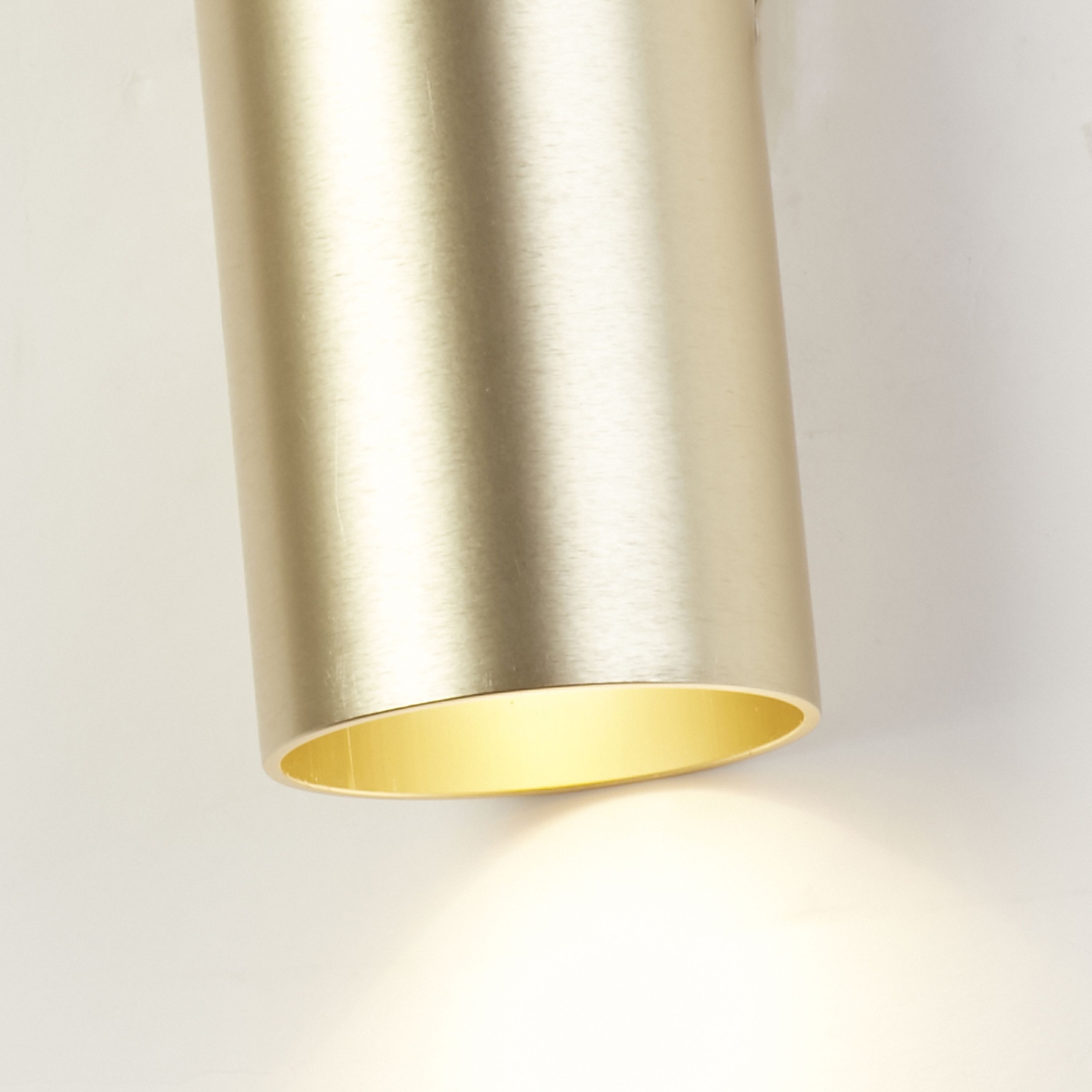 Close up of the Searchlight Cyls wall light in brushed gold showing the metal texture and lit lower aperture against a white wall