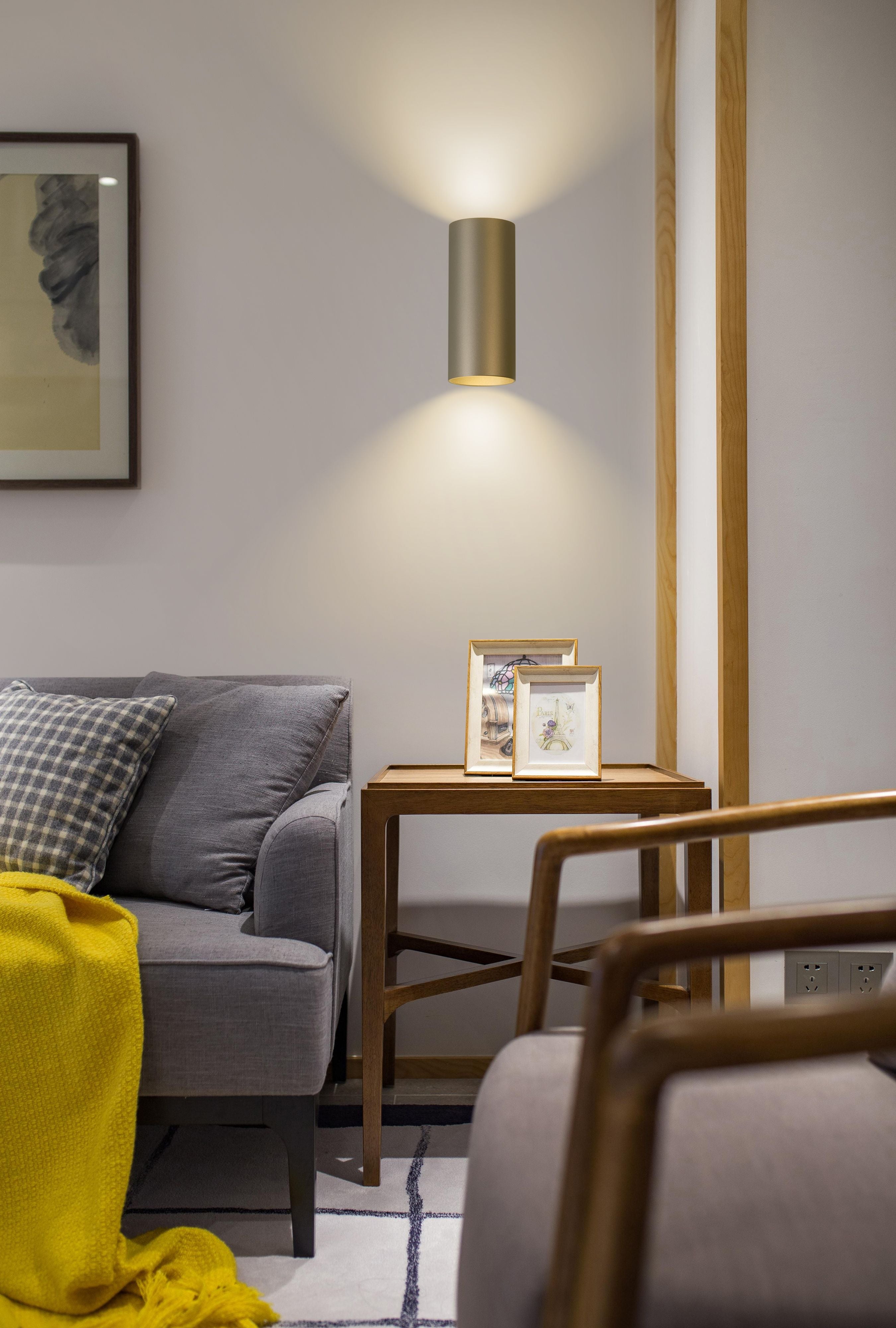 Searchlight Cyls wall light in latte finish lit up and down on a living room wall above a wooden side table with picture frames