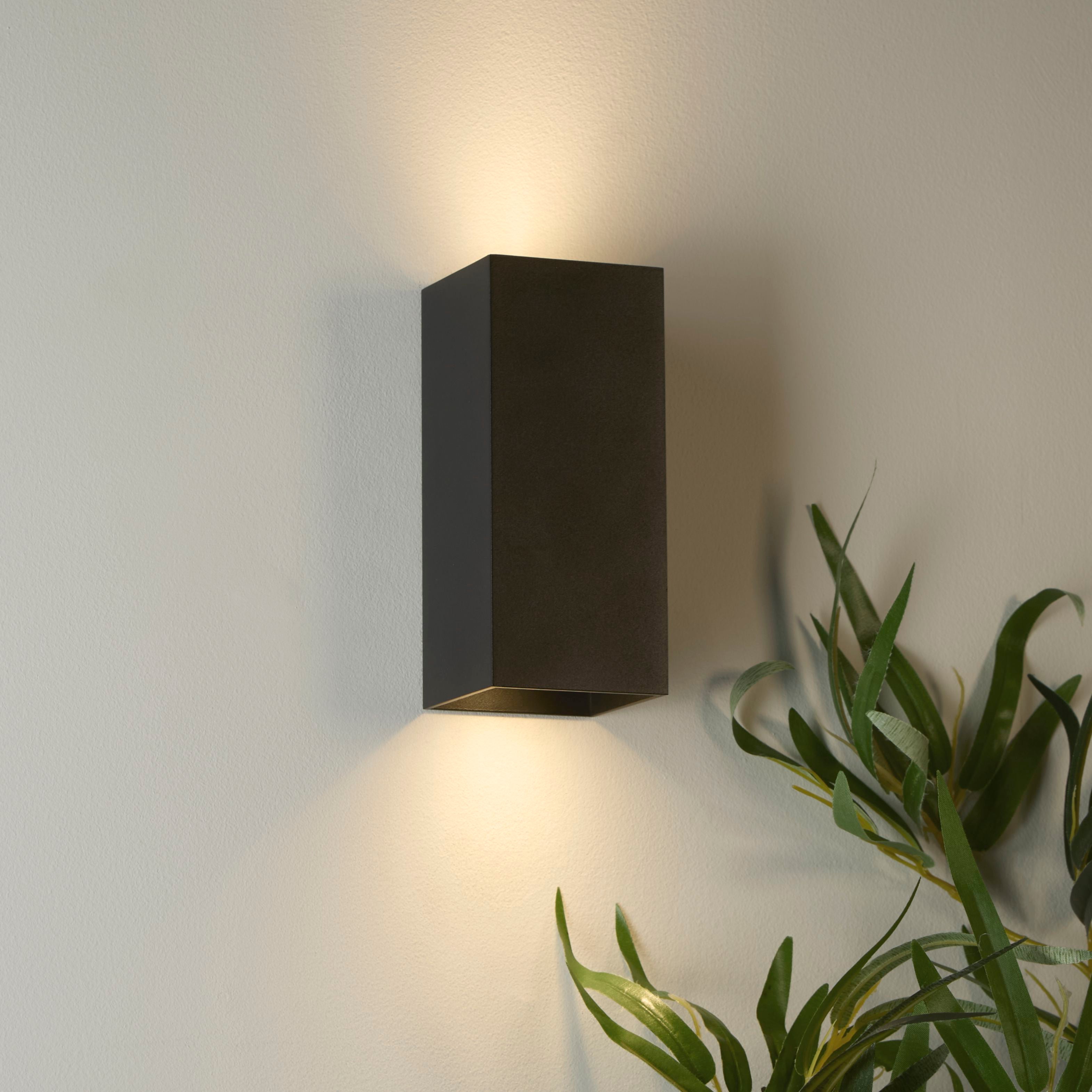 Searchlight Don wall light in matte black installed on a wall beside a plant, lit and casting light both upward and downward