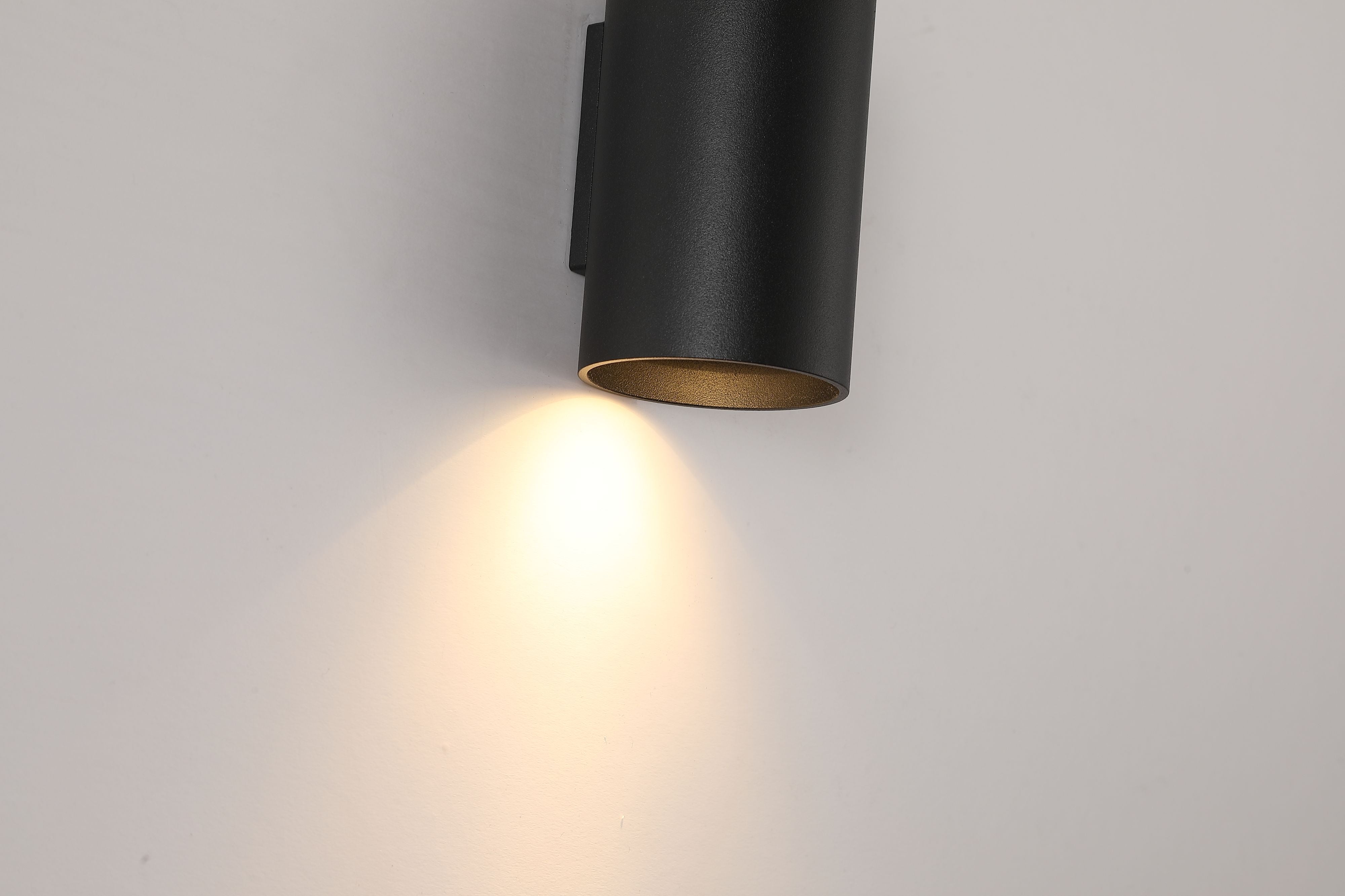 Close up of the Searchlight Cyls Cylinder Down Wall Light in black, lit, showing the warm gold interior of the shade and the light beam falling on the wall