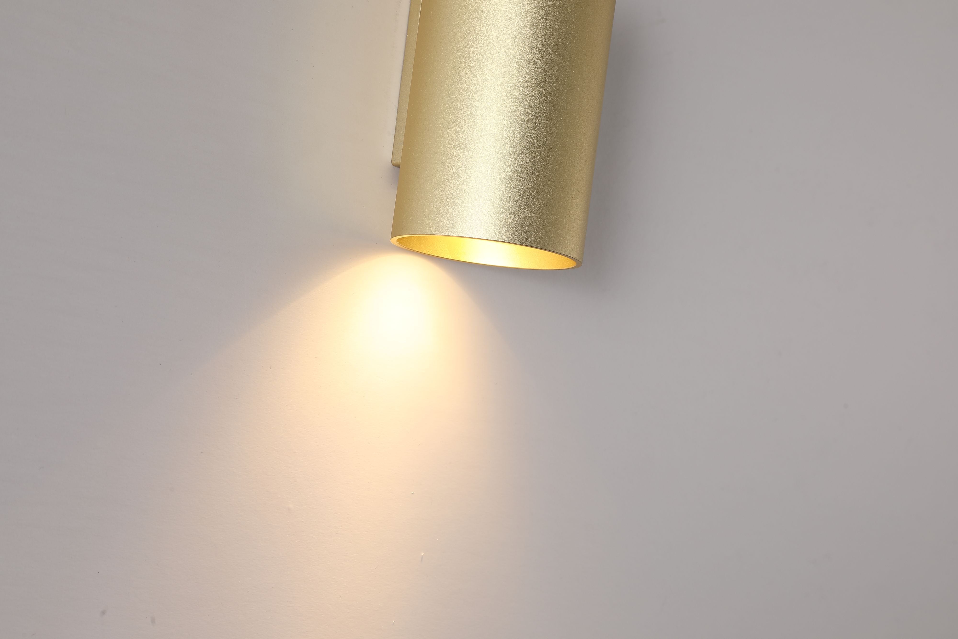 Close up of the Searchlight Cyls Cylinder Down Wall Light in gold, lit, showing the wall bracket, cylinder profile and warm light spilling onto the wall