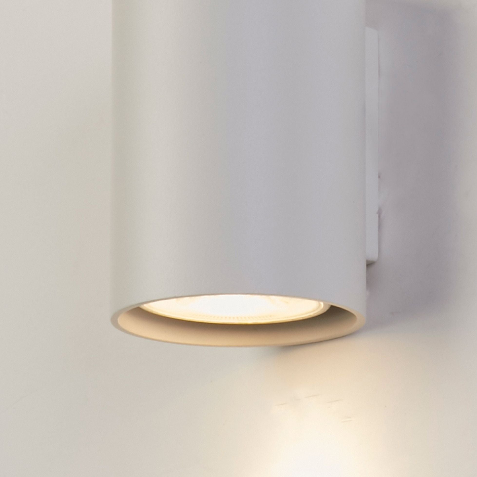 Searchlight Cyls wall light in white mounted on a plain wall, lit and casting a warm downward pool of light onto the wall below