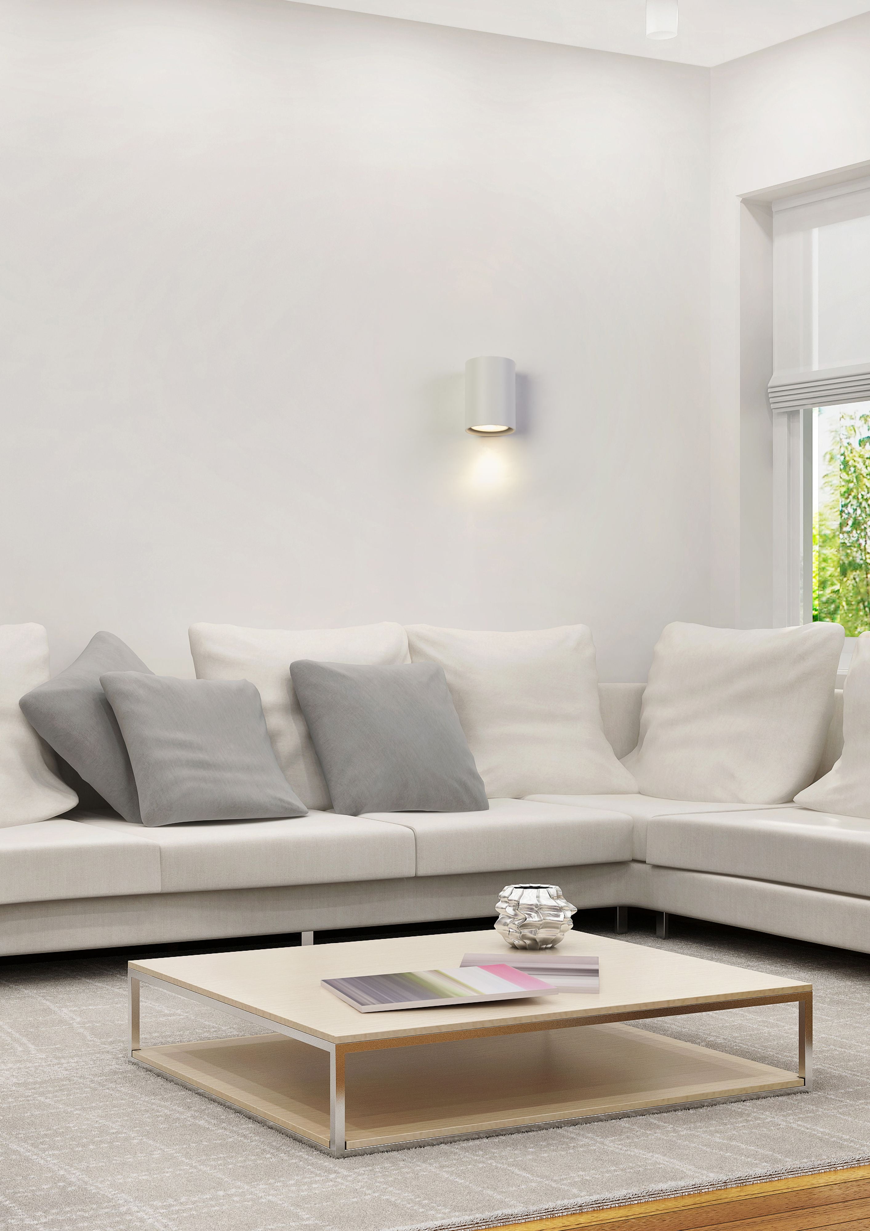 Searchlight Cyls wall light in white shown lit above a light grey corner sofa in a living room, casting a warm downward glow