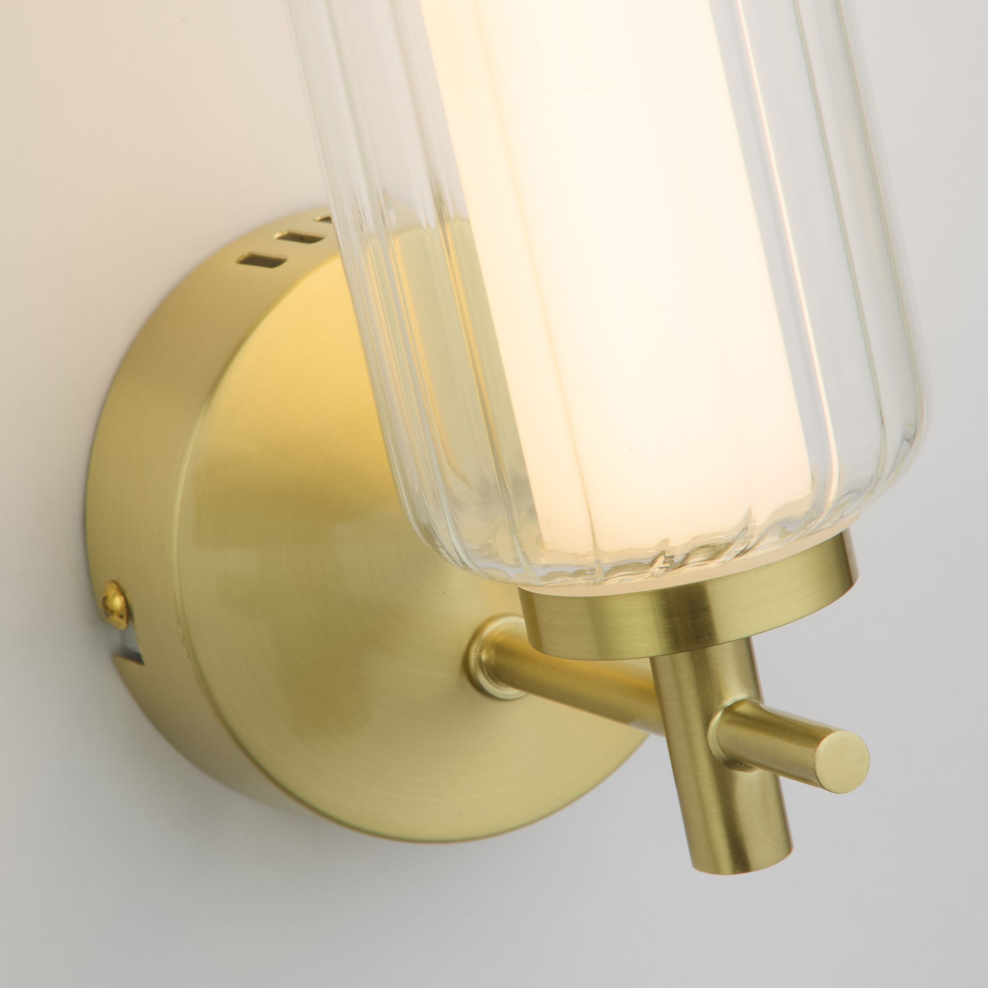 Close up of the satin brass backplate, arm and toggle switch on the Mod wall light, ribbed glass shade above