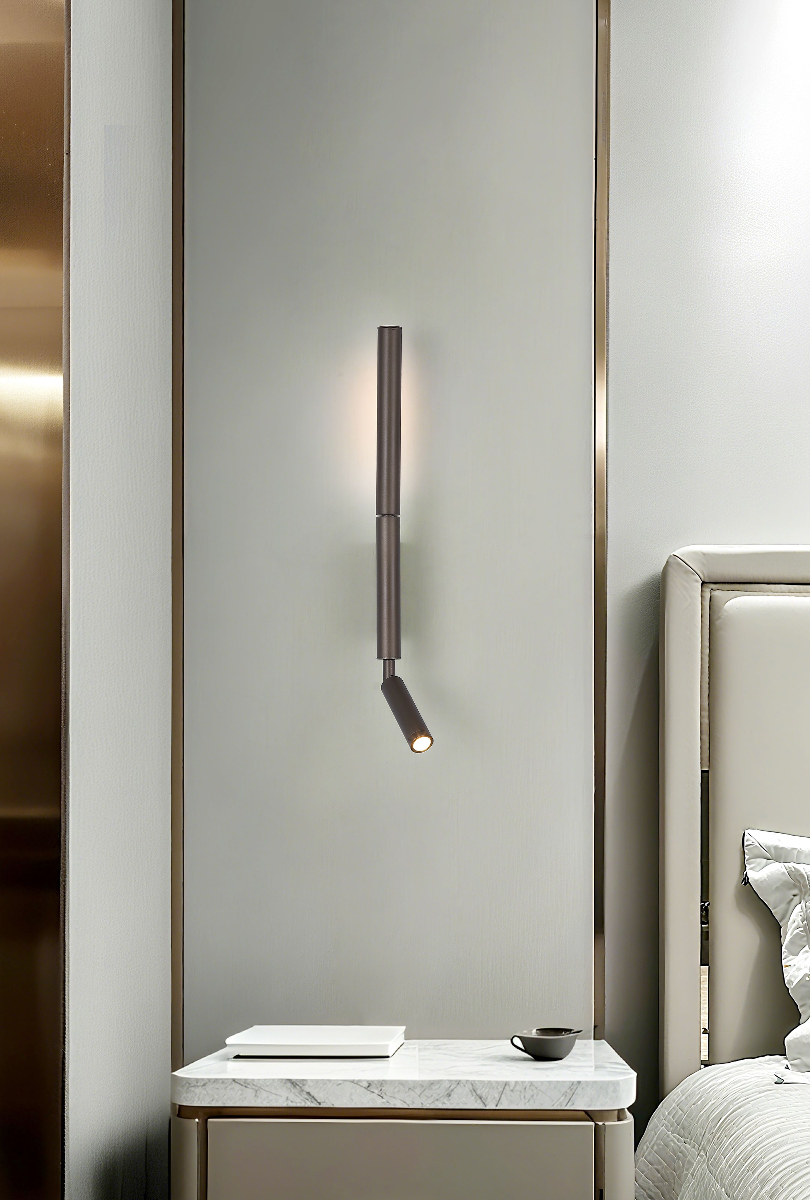 Searchlight Architect LED Wall Light with Reading Arm in latte metal, mounted above a marble topped bedside table with books and a cup, warm ambient glow up the wall and the reading light angled down