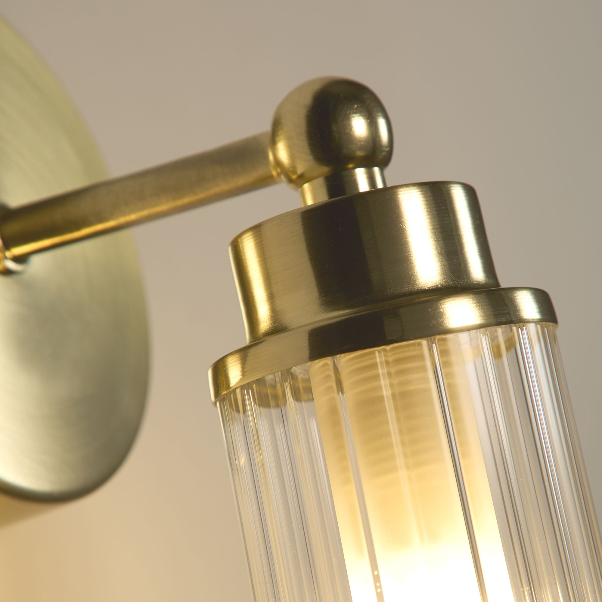 Close up of the satin brass gimbal fitting and ribbed glass shade on the Searchlight Imperial wall light, bulb lit