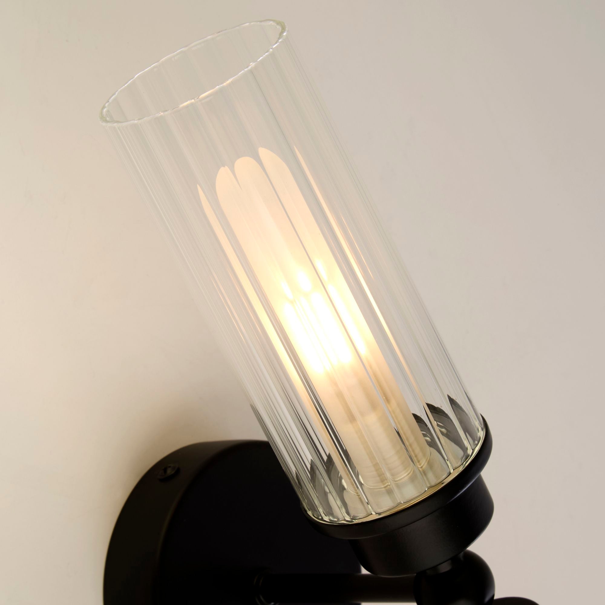 Close up at an angle of one matt black gimbal fitting and ribbed glass shade on the Searchlight Imperial 2 light wall light, bulb lit