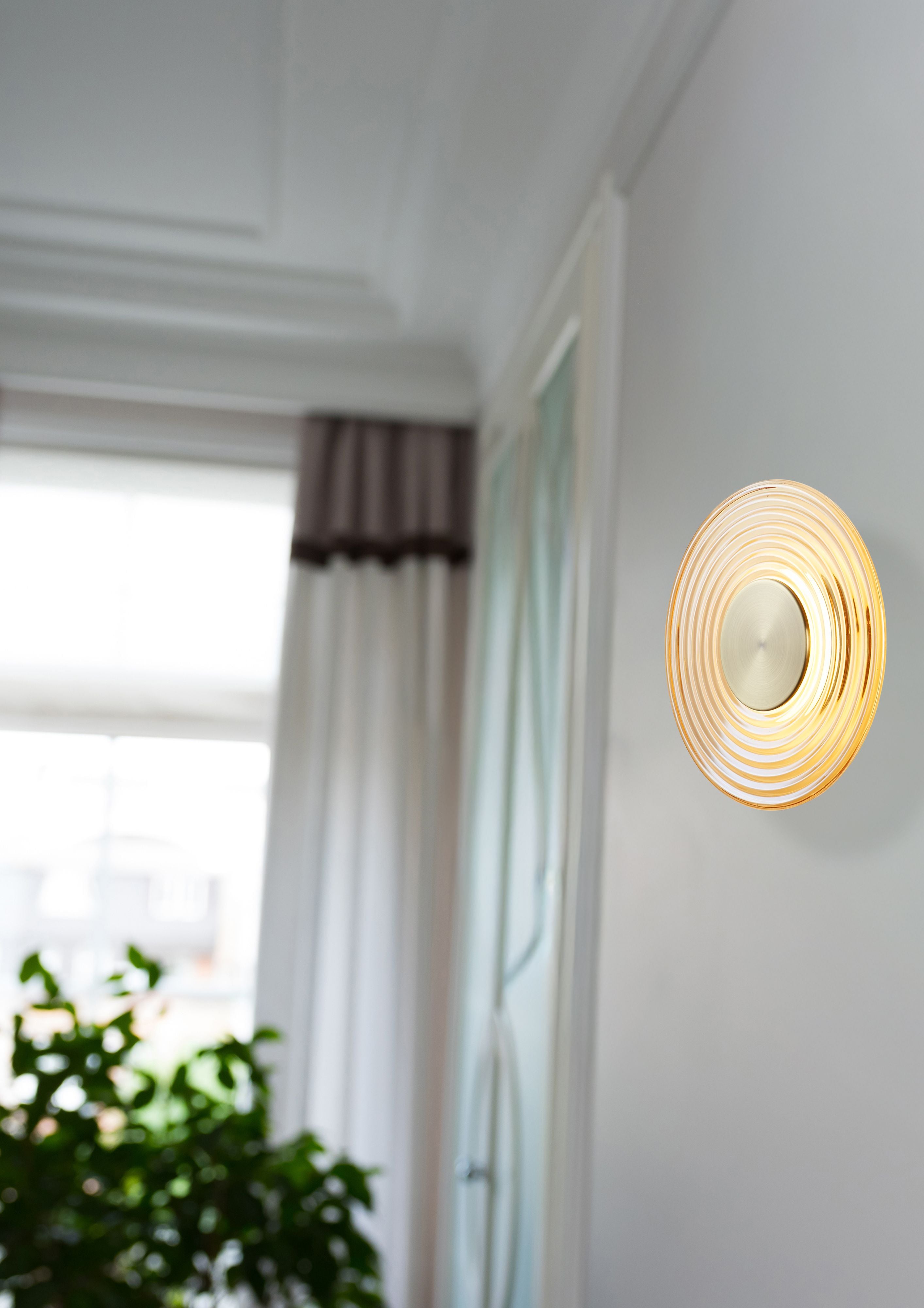 Hallway with a curtained window, showing the Cleo wall light in antique brass and amber glass lit and mounted on a pale wall