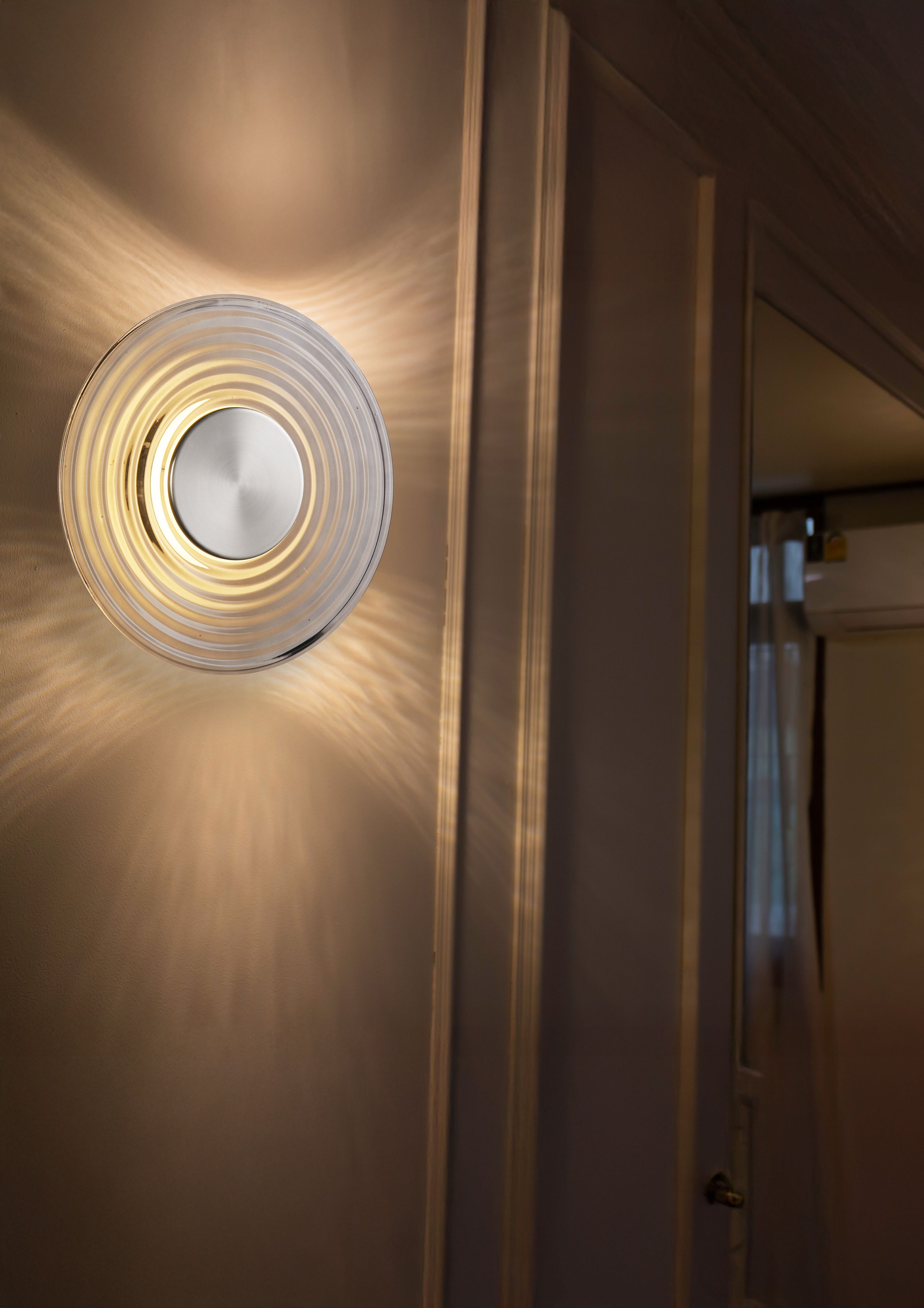 Cleo wall light lit and mounted beside a doorway, casting a warm ringed glow across the wall