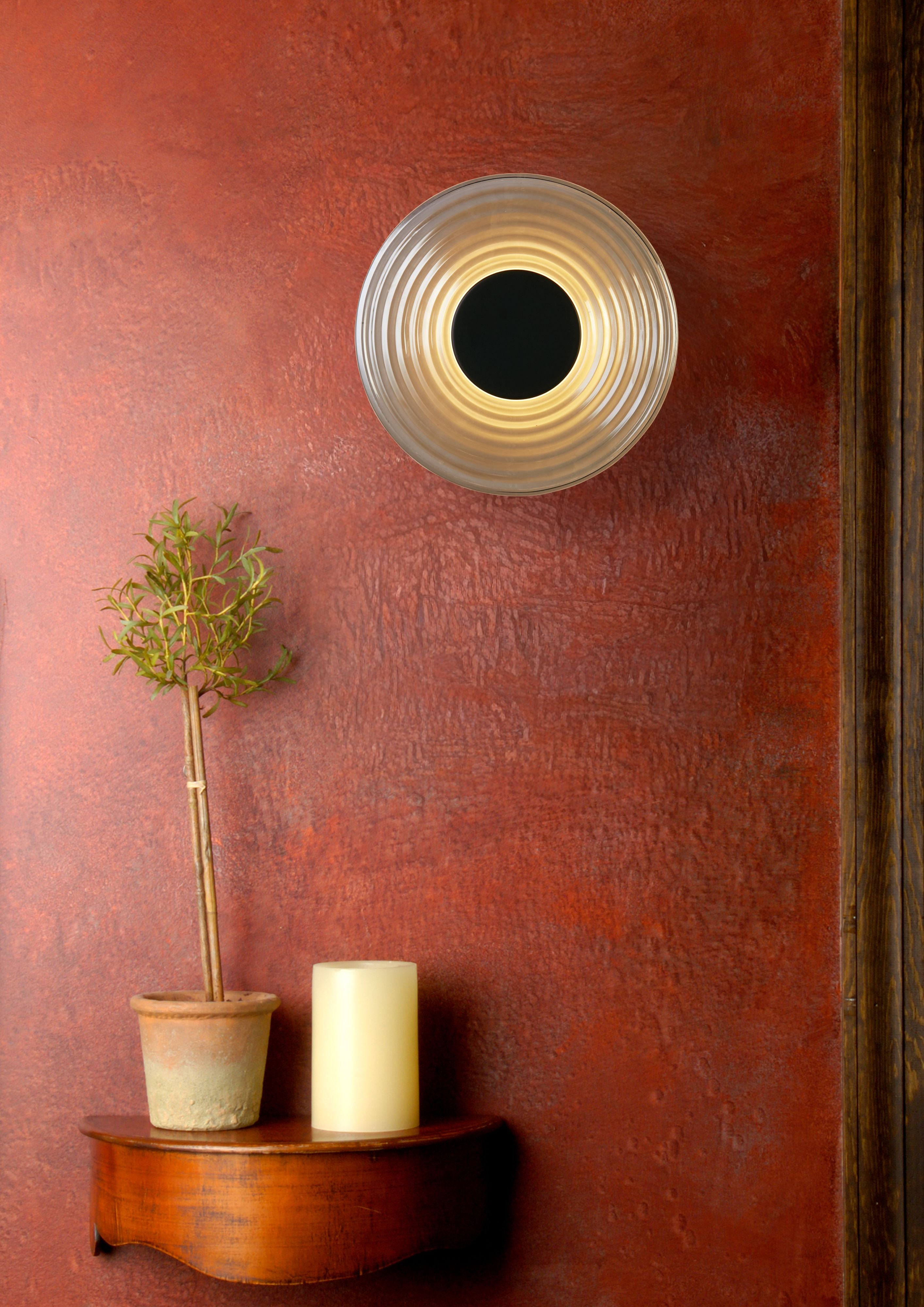 Cleo wall light lit and mounted on a terracotta coloured wall beside a small potted olive tree and candle