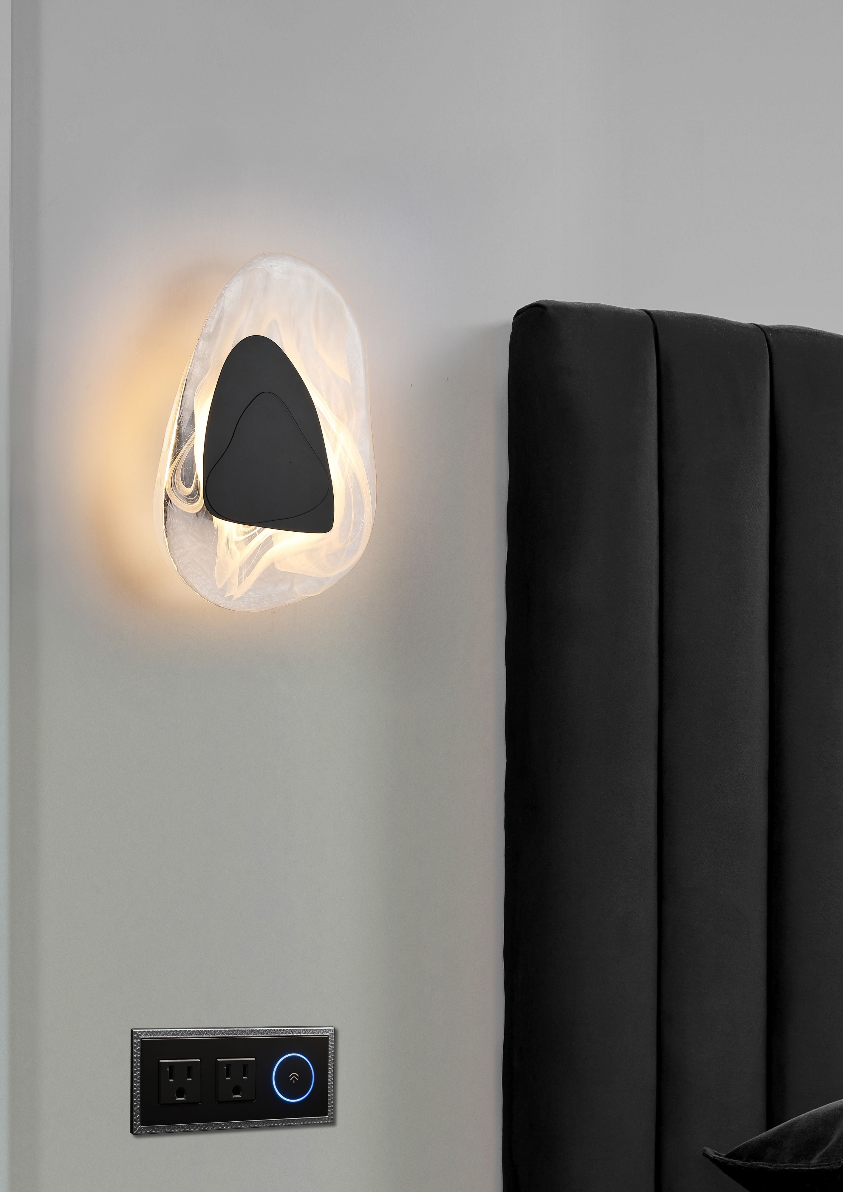 Destiny LED wall light in matt black installed above a bed, glowing beside a black upholstered headboard