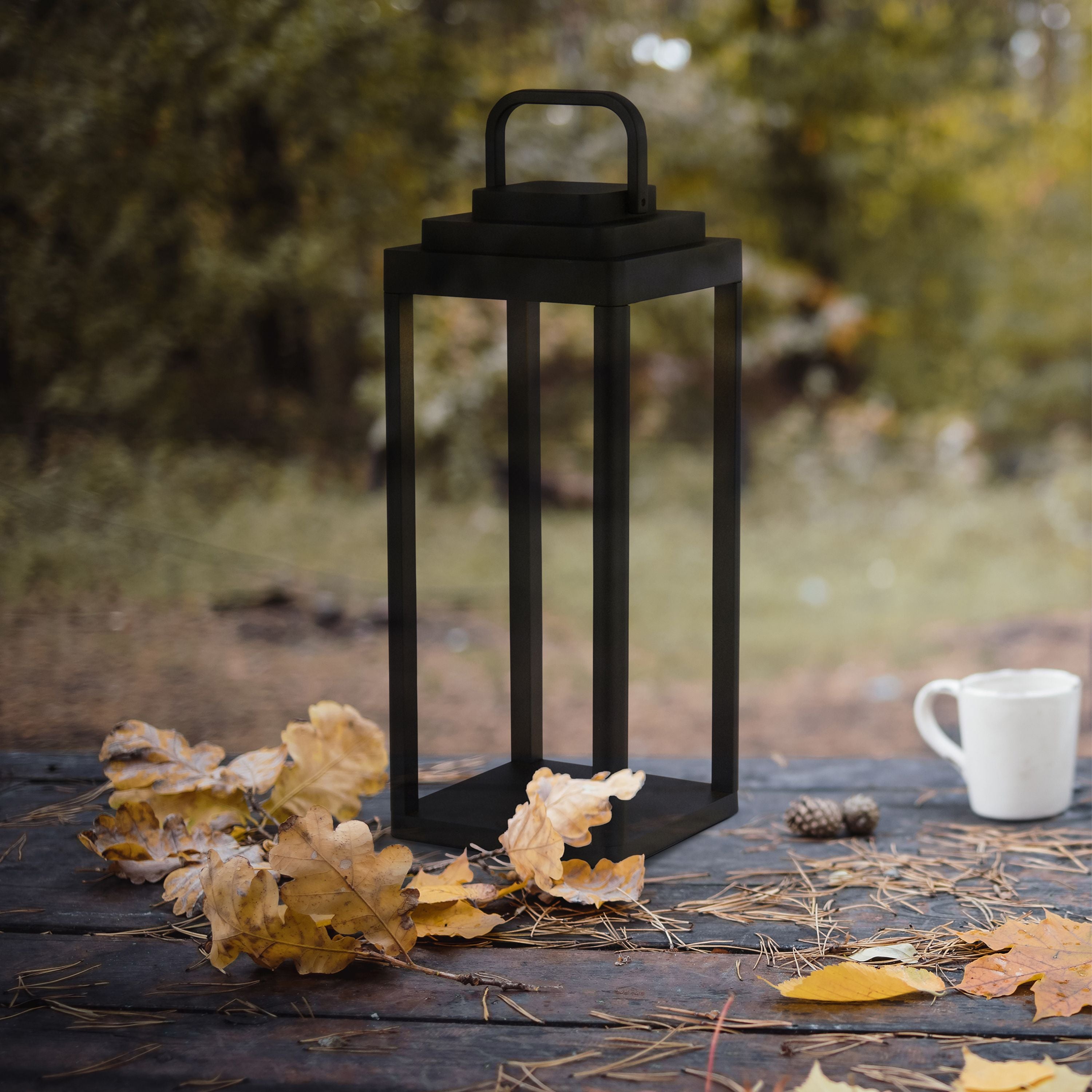 Portobello black outdoor table lamp on a wooden table surrounded by autumn leaves and a mug