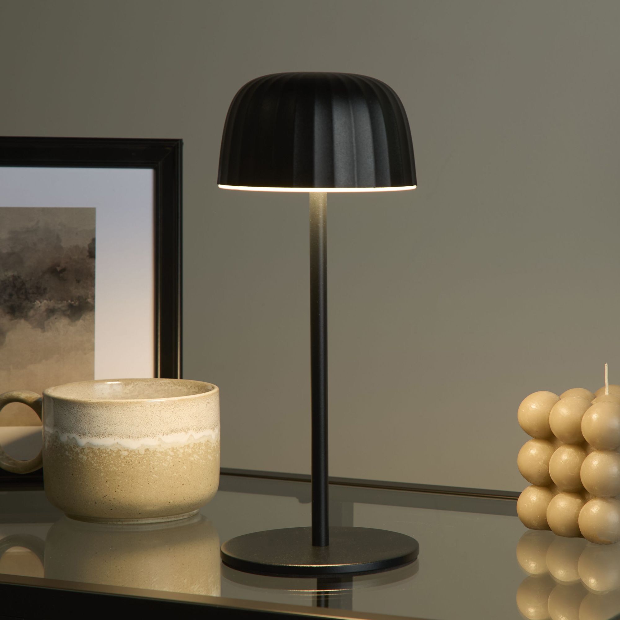 Ezra black table lamp lit on a dark glass table at night beside a ceramic mug, framed print, and a candle