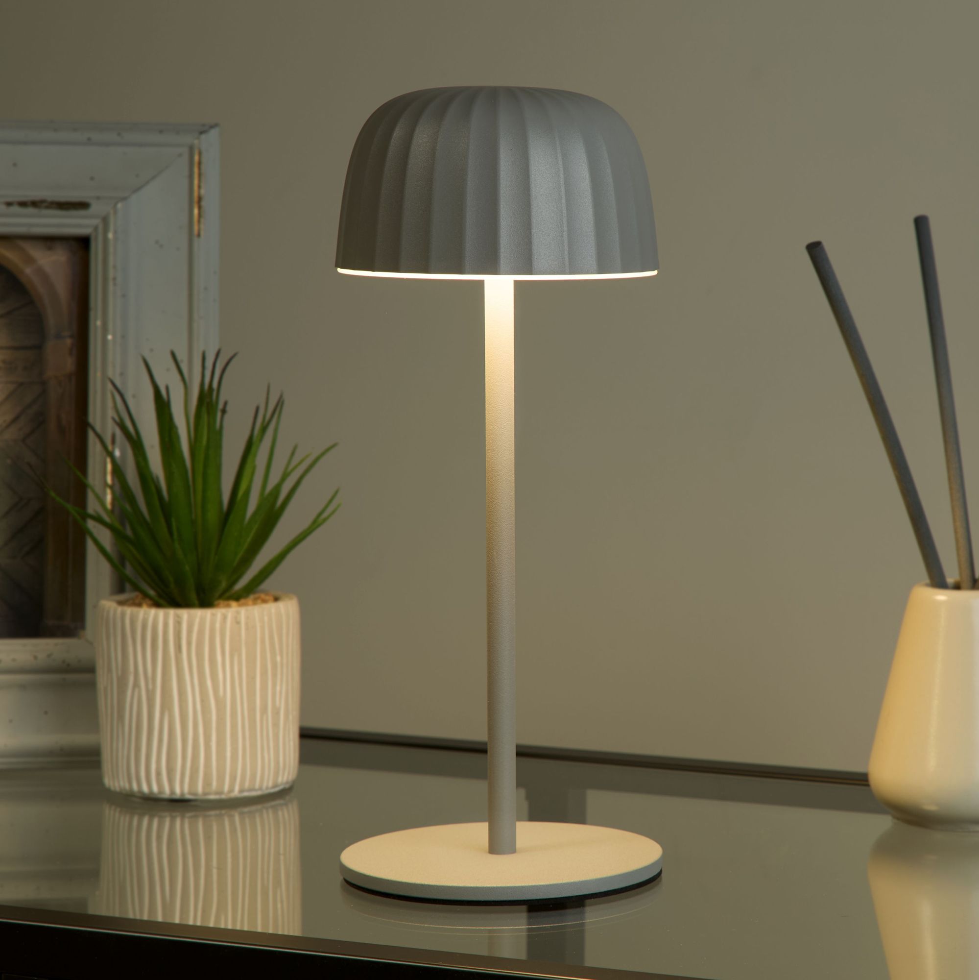 Ezra grey table lamp lit on a dark glass table at night beside a potted plant and a ceramic vase