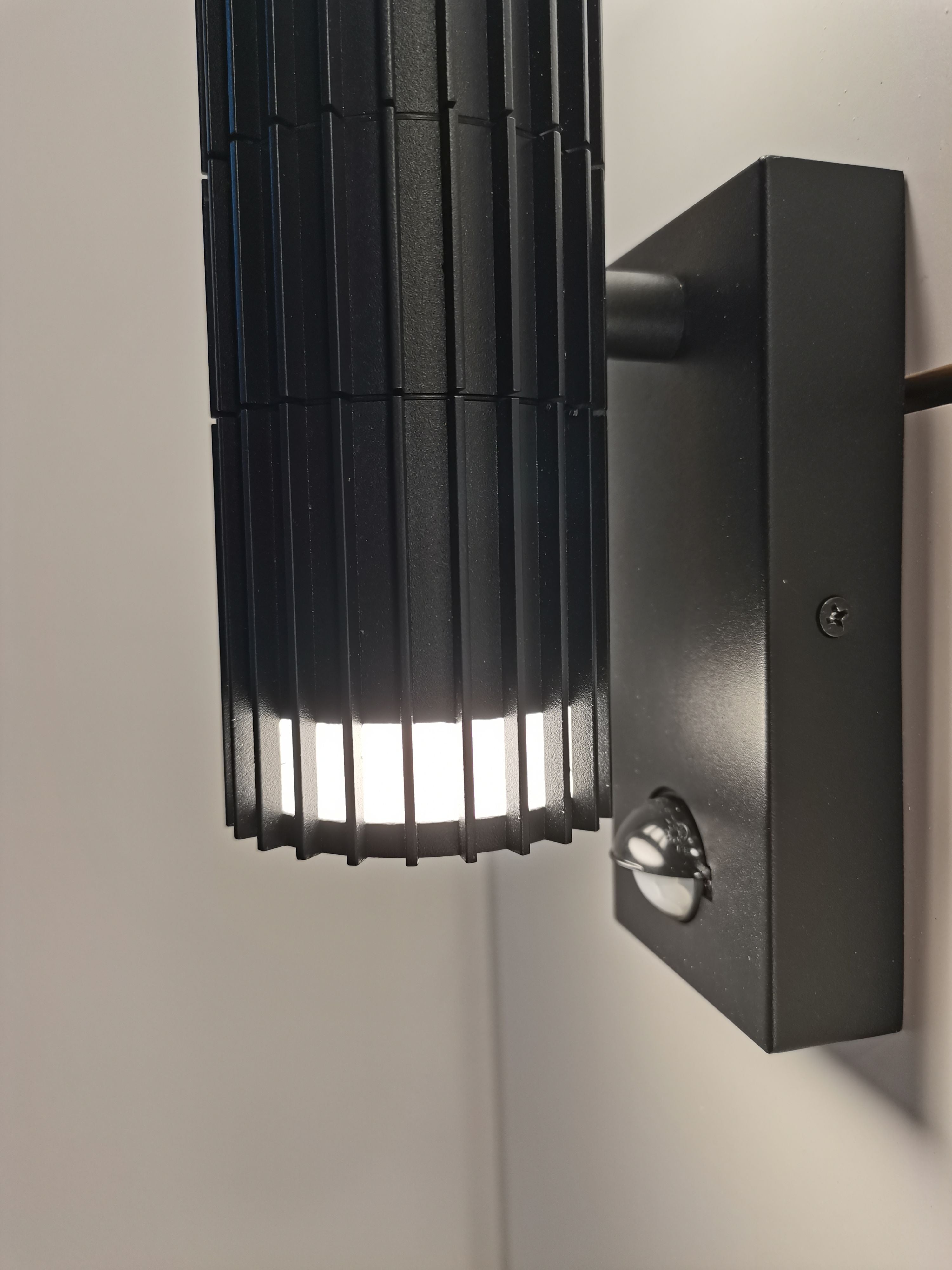 Close up angled view of the Detroit PIR black wall light mounted on a white wall, showing the round motion sensor lens on the base plate