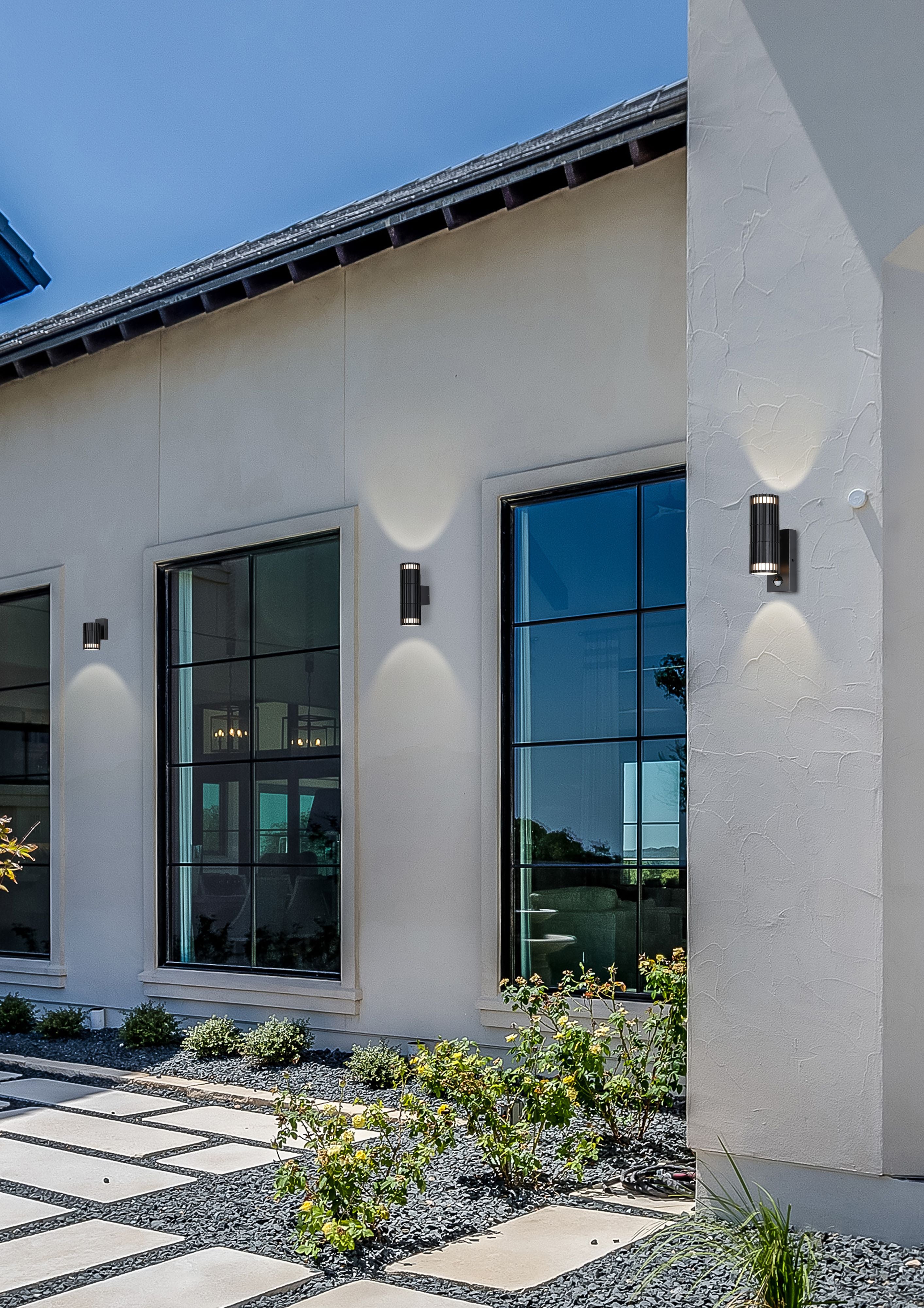 Lifestyle photo of matte black Detroit outdoor wall lights installed either side of a modern stucco house entrance, one with a visible motion sensor, glowing up and down against the wall with landscaped planting and paving in the foreground