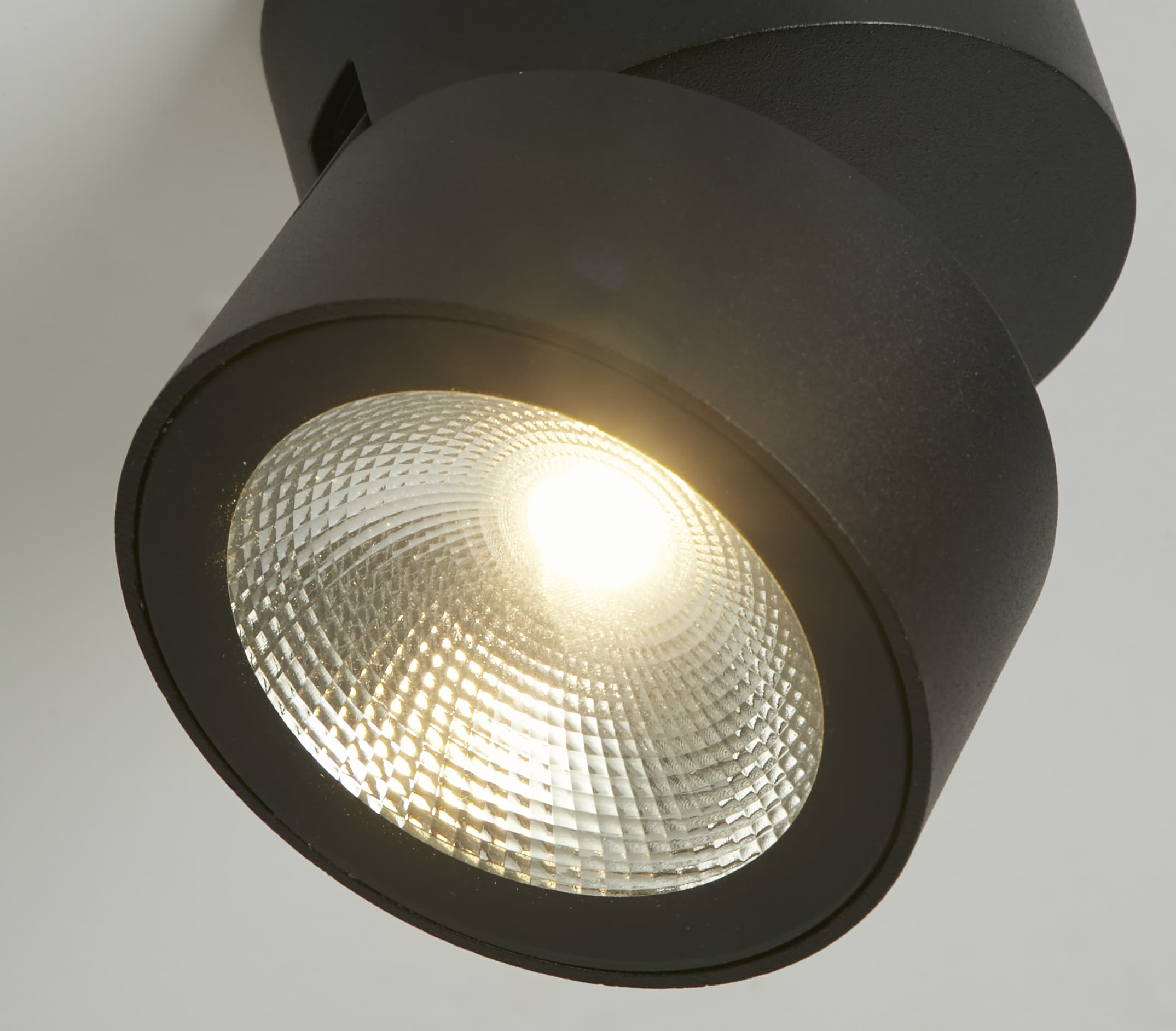 Extreme close up of the Manhattan spotlight's lens and warm LED glow, showing the swivel joint between the round base and the light head