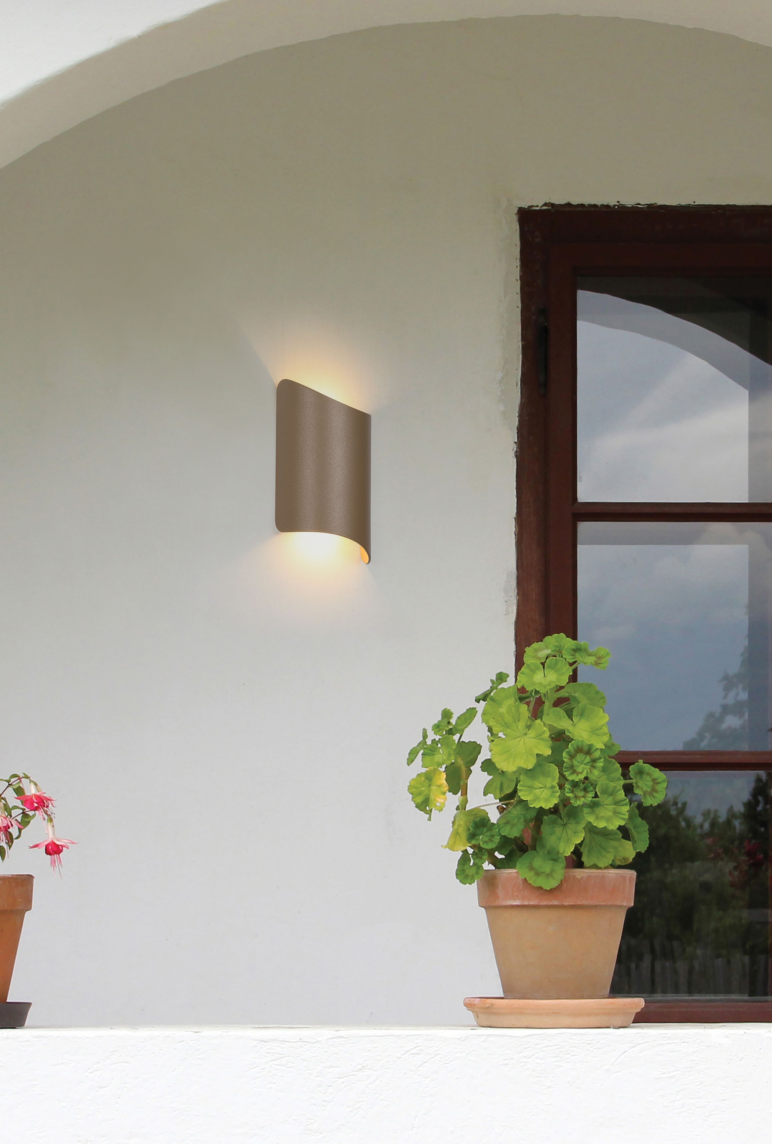 Shard outdoor wall light in gold installed beside a window on a rendered exterior wall, lit, with potted plants below