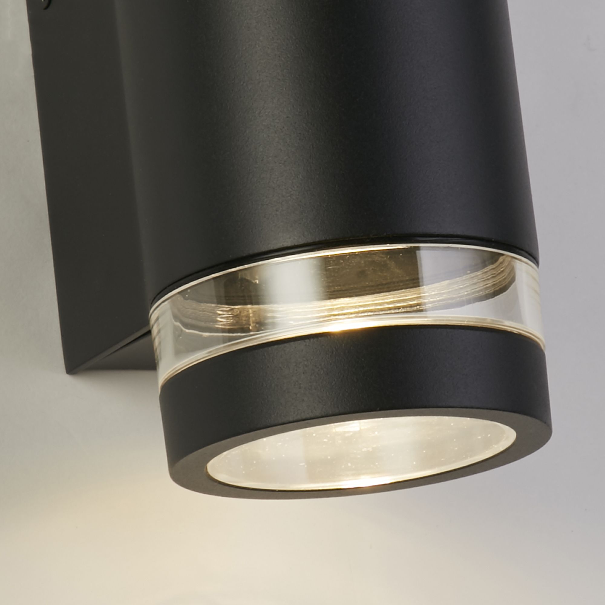 Close up angled view of the illuminated lower lens ring on the Austin black wall light, mounted on a white wall