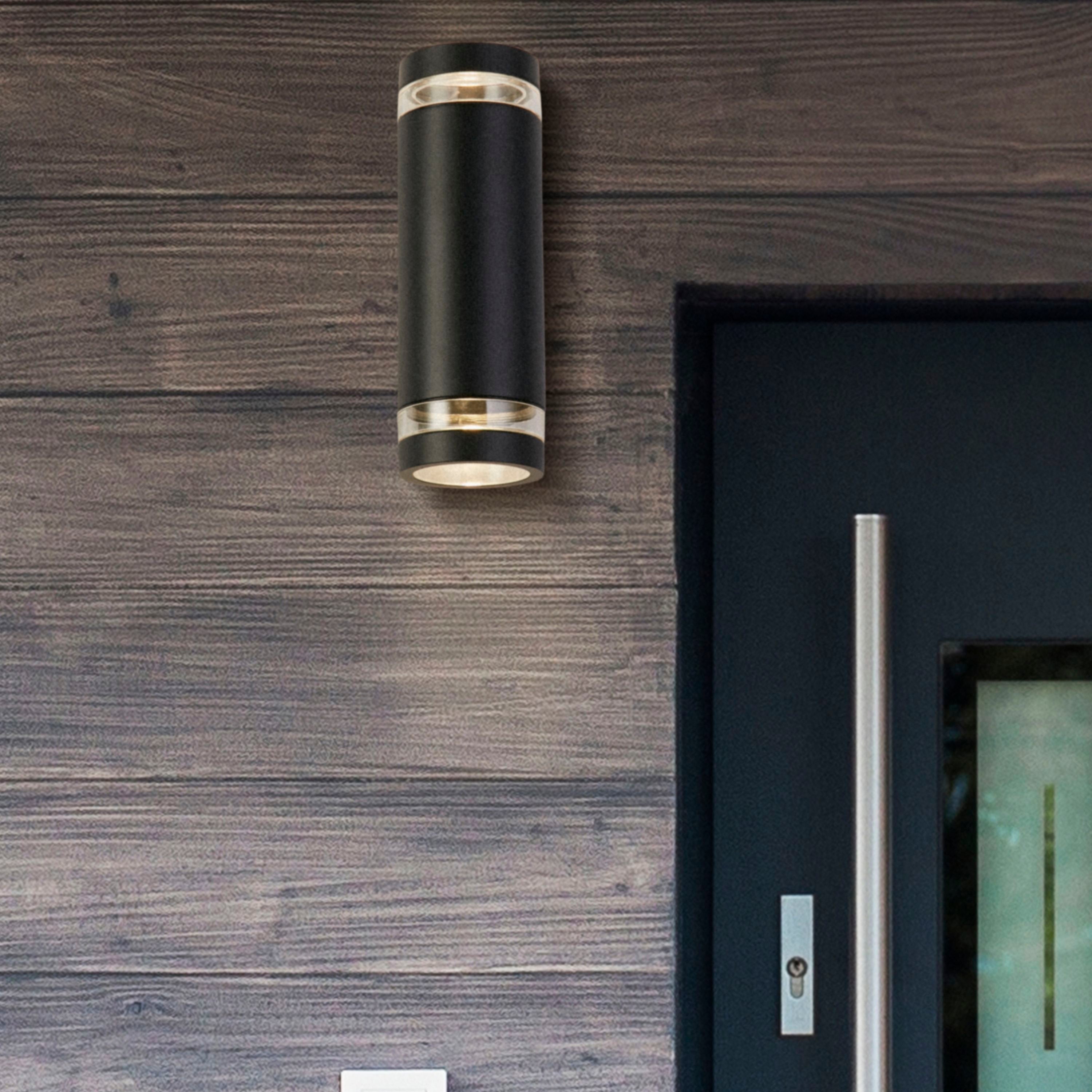 Lifestyle photo of the Austin black wall light mounted on a dark wood clad wall beside a front door, glowing warm light up and down