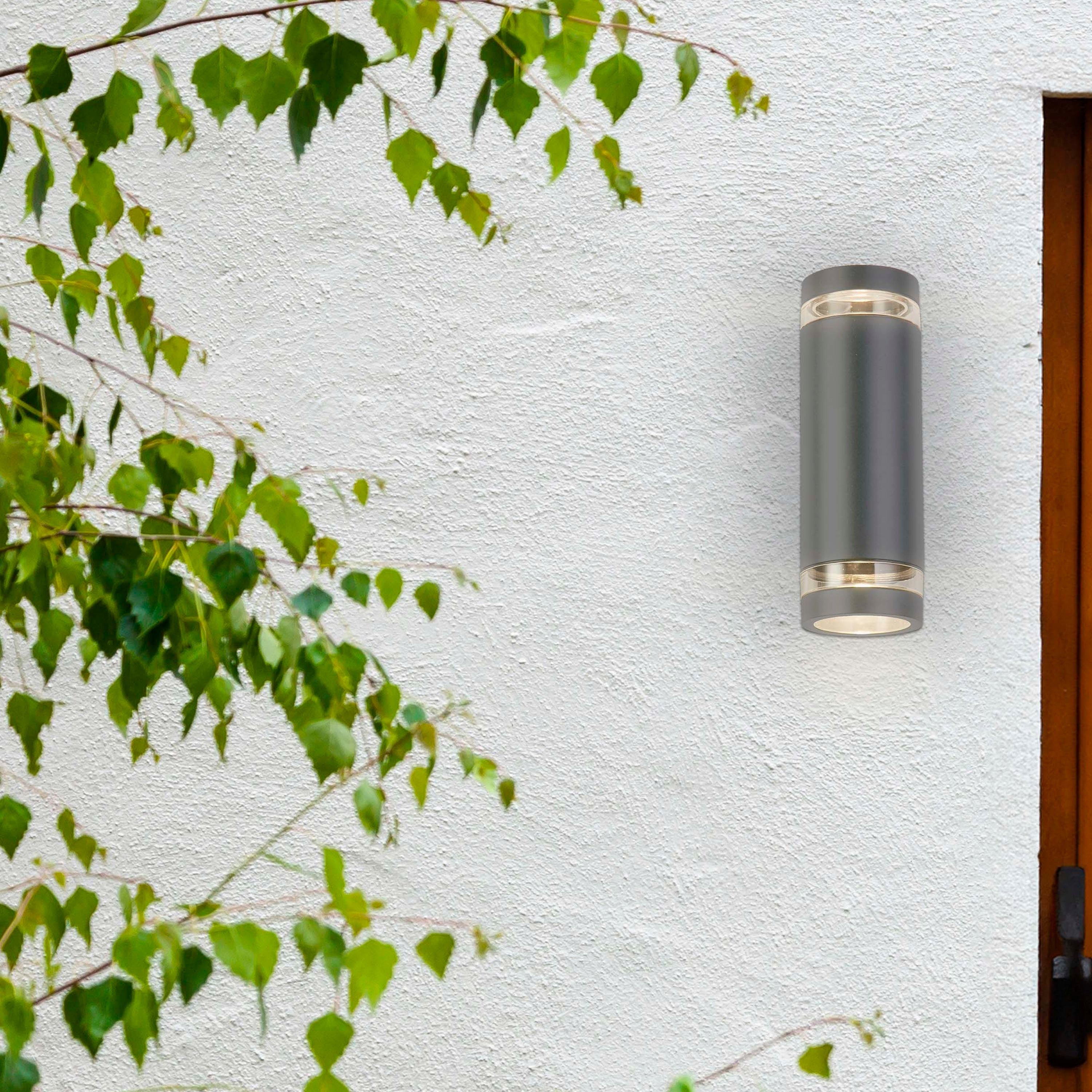 Lifestyle photo of the Austin grey wall light mounted on a white textured exterior wall beside a wooden door frame, with green foliage in the foreground, glowing warm light up and down