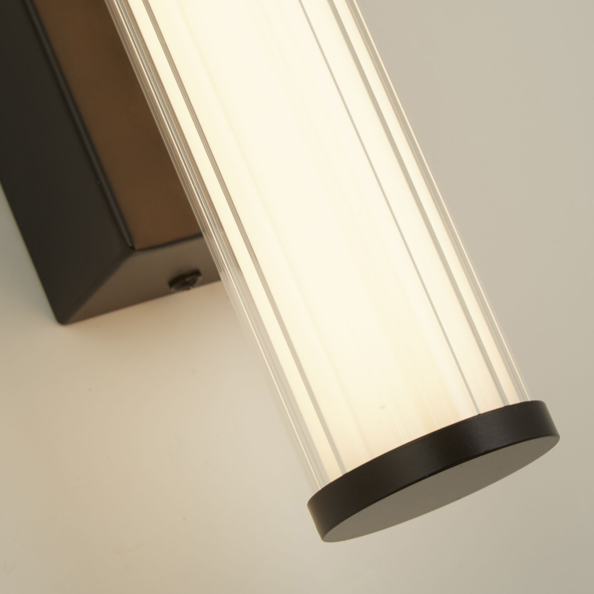 Close up of the black clamp bracket and ribbed glass tube on the Searchlight Clamp wall light, lit