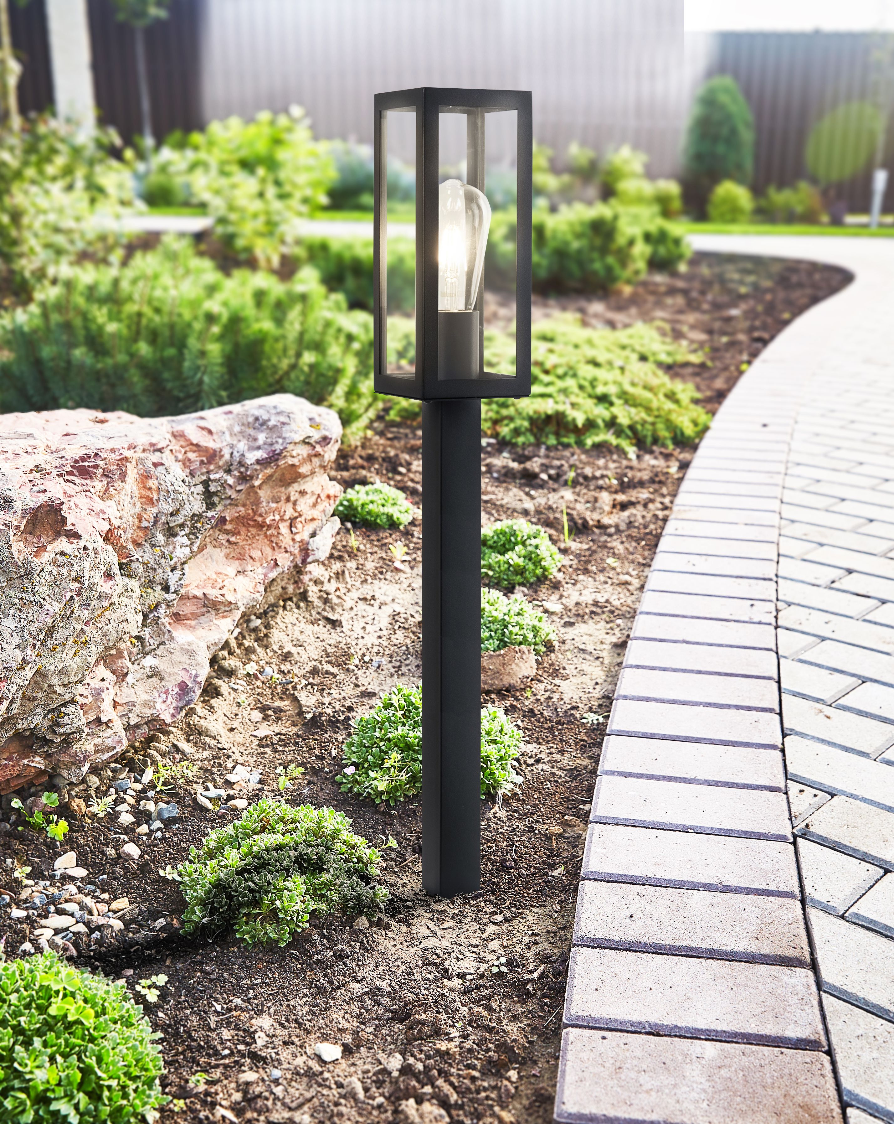 Box outdoor post light installed in a garden border beside a stone and paved path, lit at dusk with the bulb glowing inside the glazed head